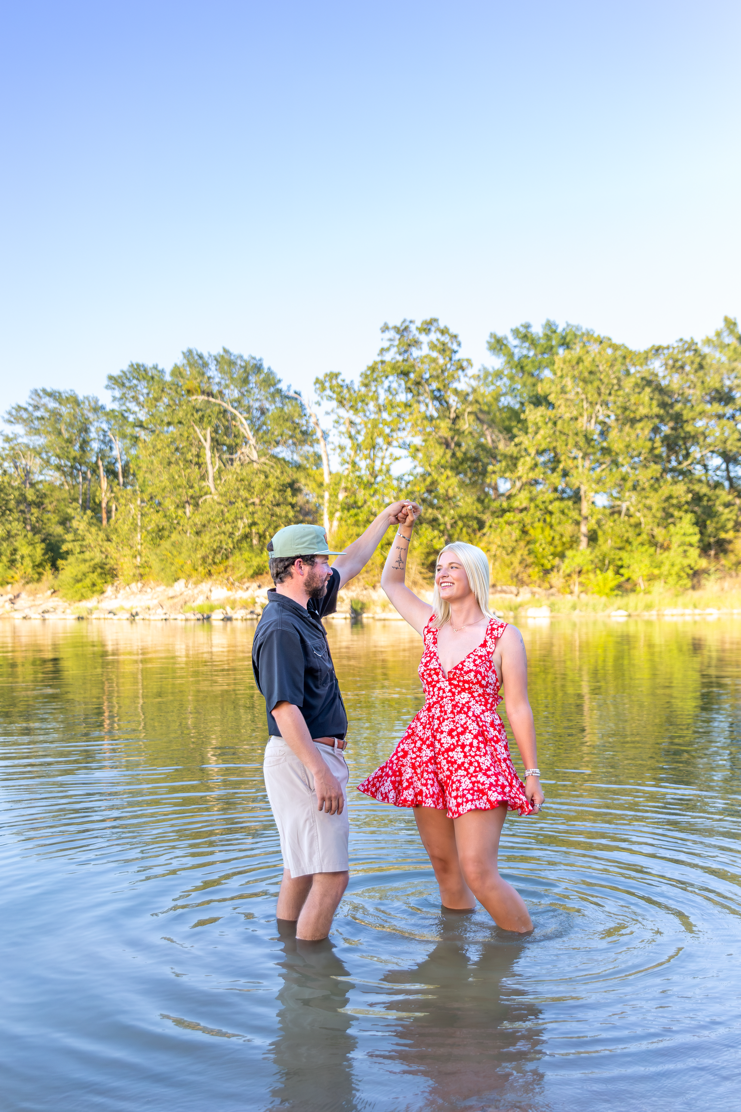 Couple dancing in shallow river water, woman in red floral dress, man in green hat, surrounded by lush trees on sunny day.