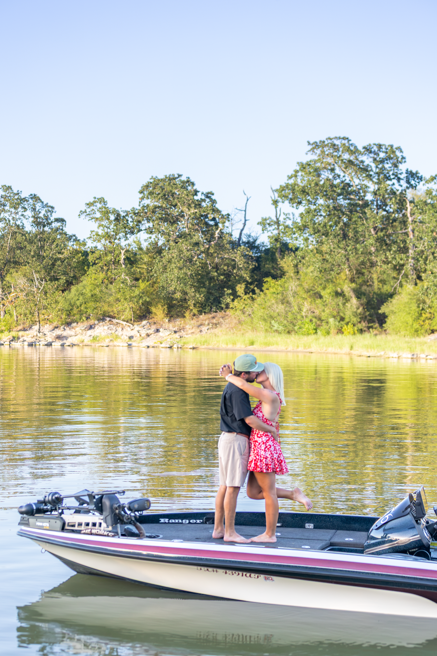 A couple shares a romantic kiss while standing on a bass boat on a calm river surrounded by lush green trees.