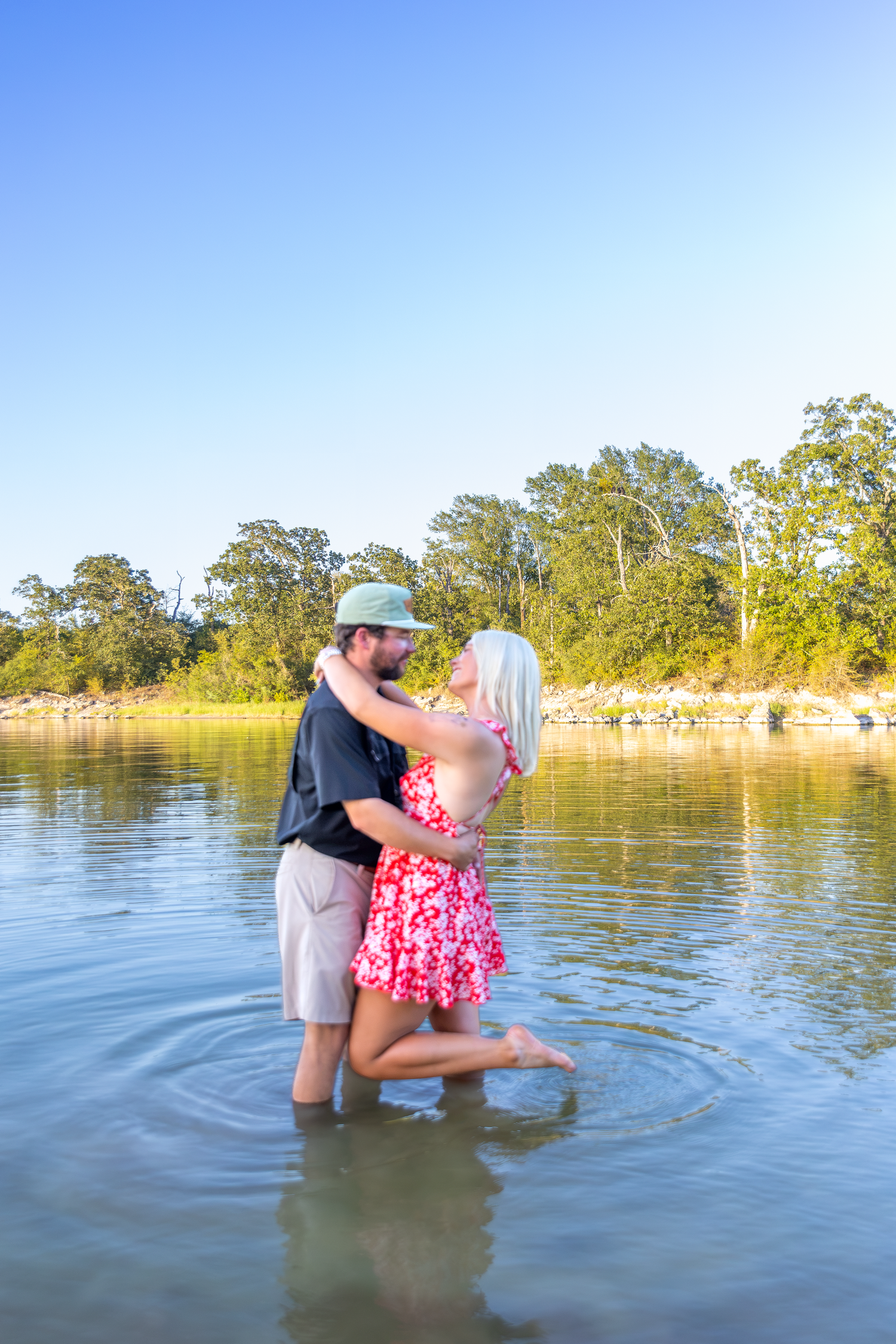 Couple embracing romantically while standing in a shallow river, surrounded by lush green trees under a clear blue sky.