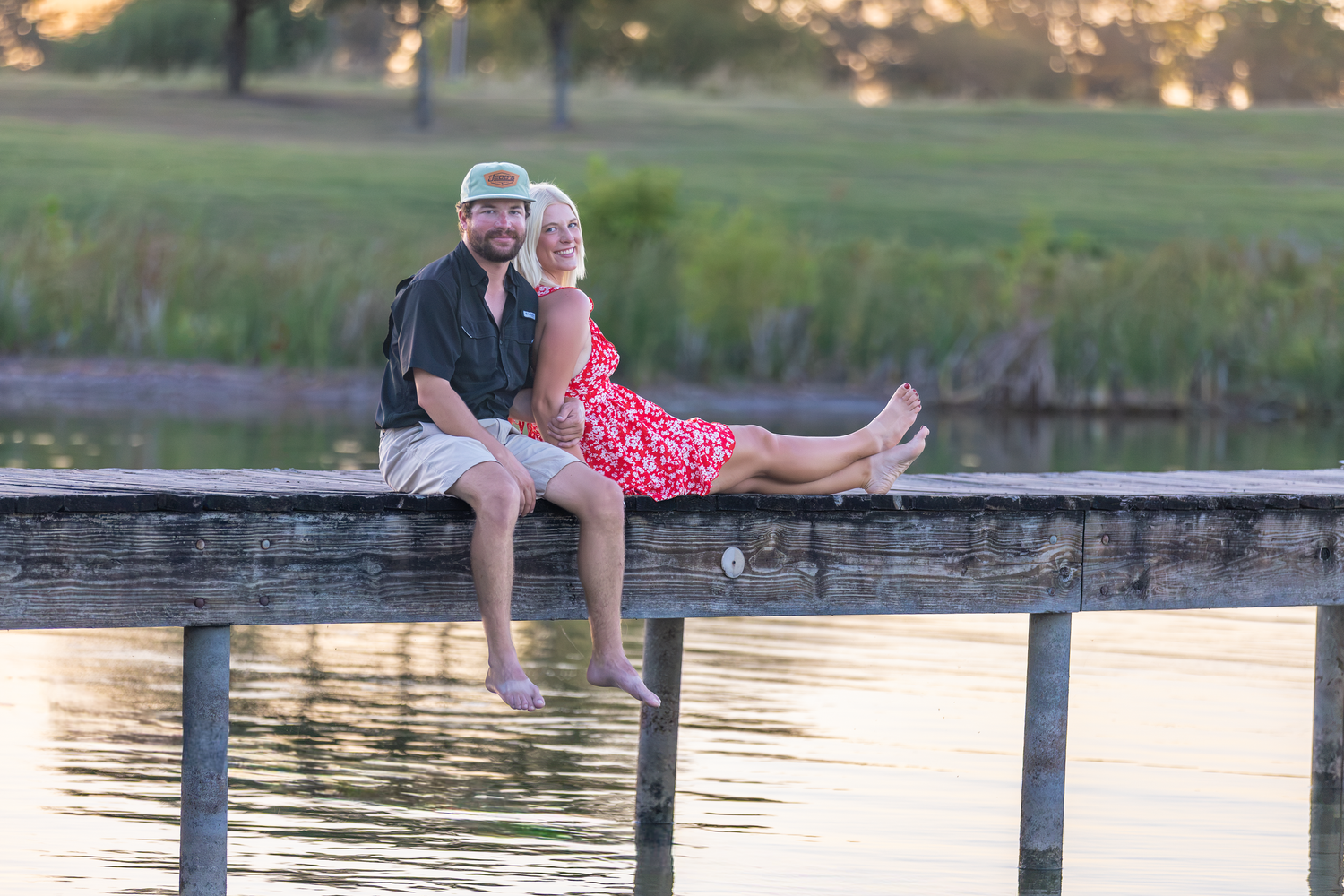 Couple sitting together on a wooden dock by a calm lake at sunset, woman in red dress leaning on man.