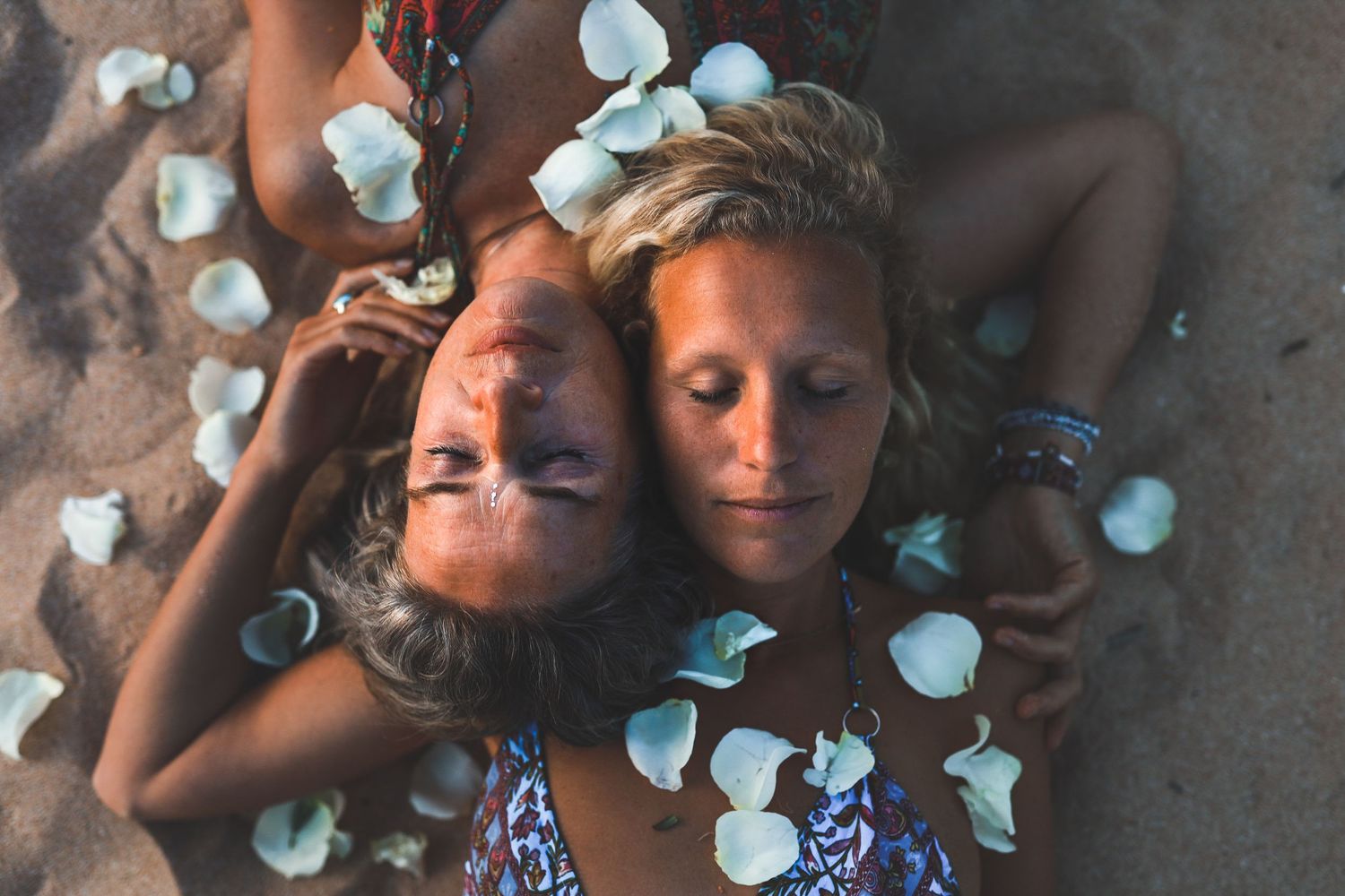 Two young women lying on sand surrounded by white flower petals, eyes closed, heads together.