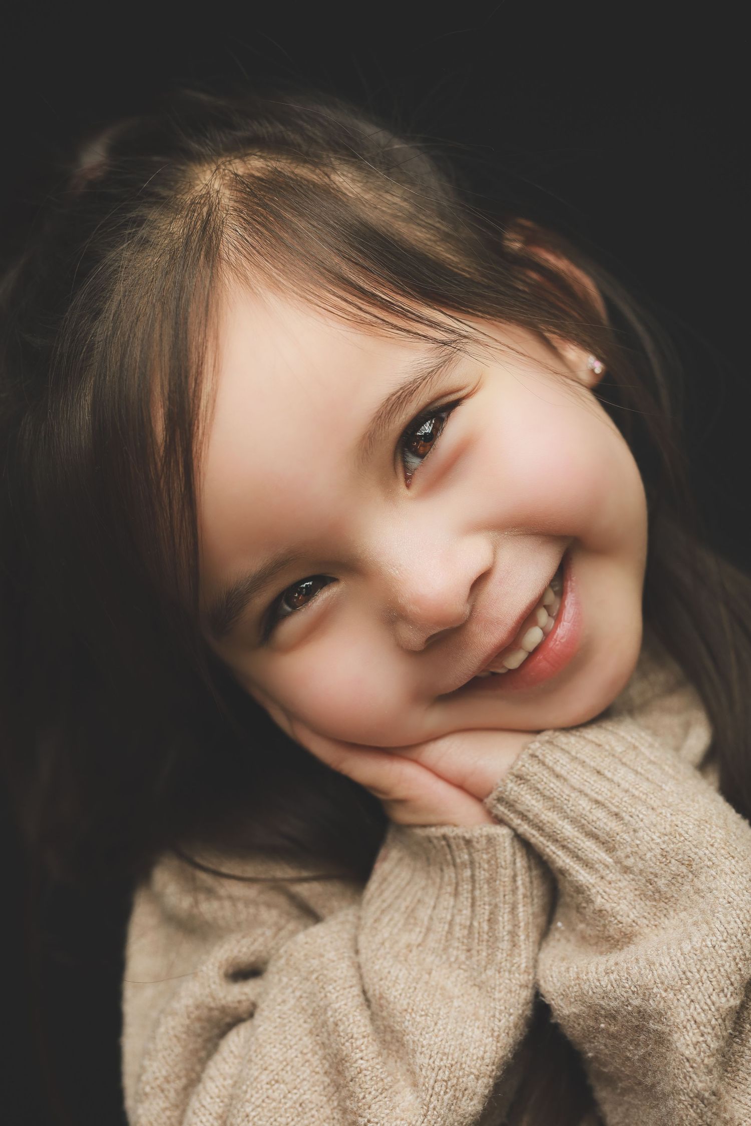 A smiling young girl with brown hair rests her chin on her hands, wearing a beige knit sweater against a dark background.