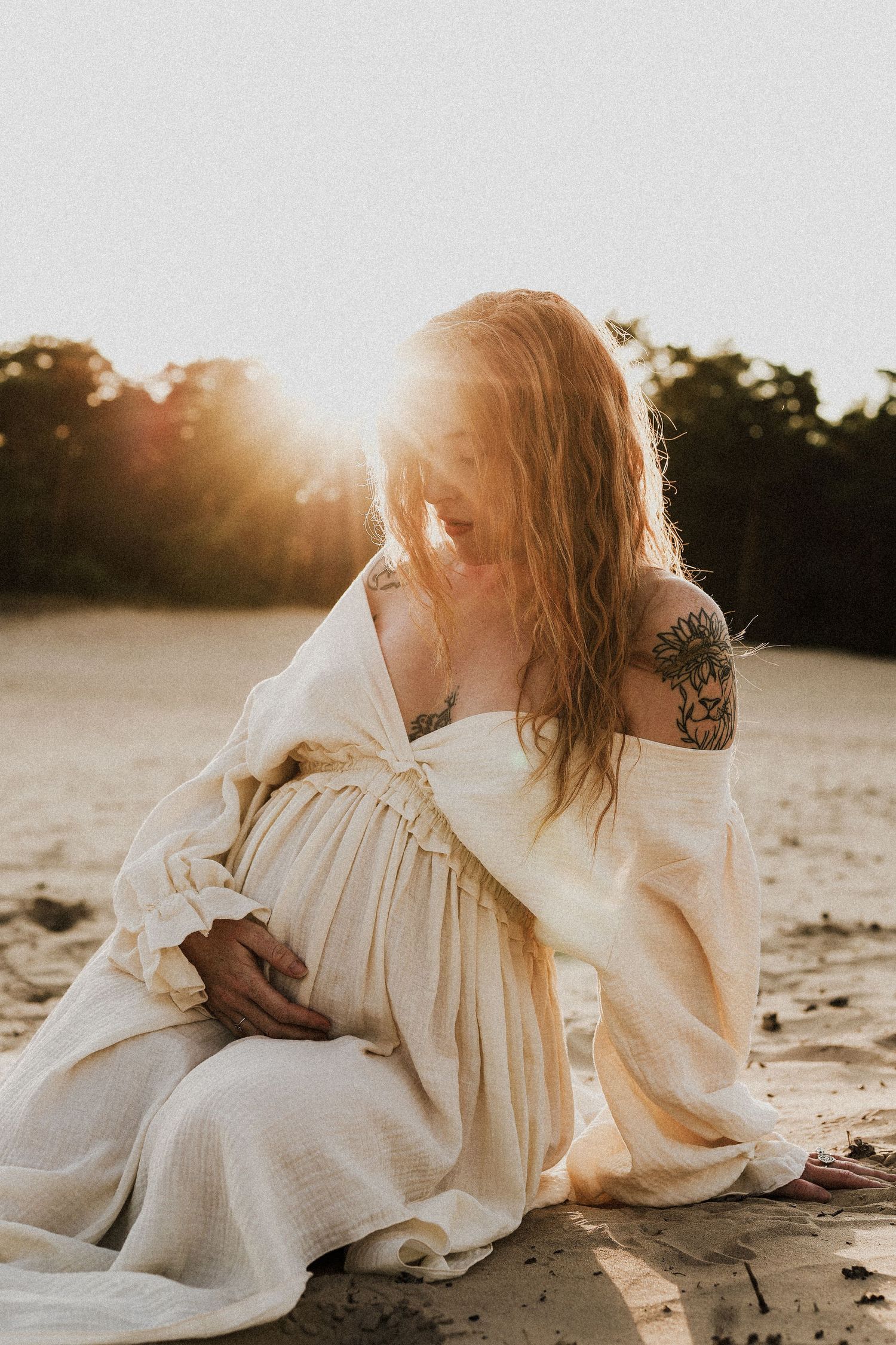 Pregnant woman with tattoos in white dress sitting on sand bathed in golden sunset light.