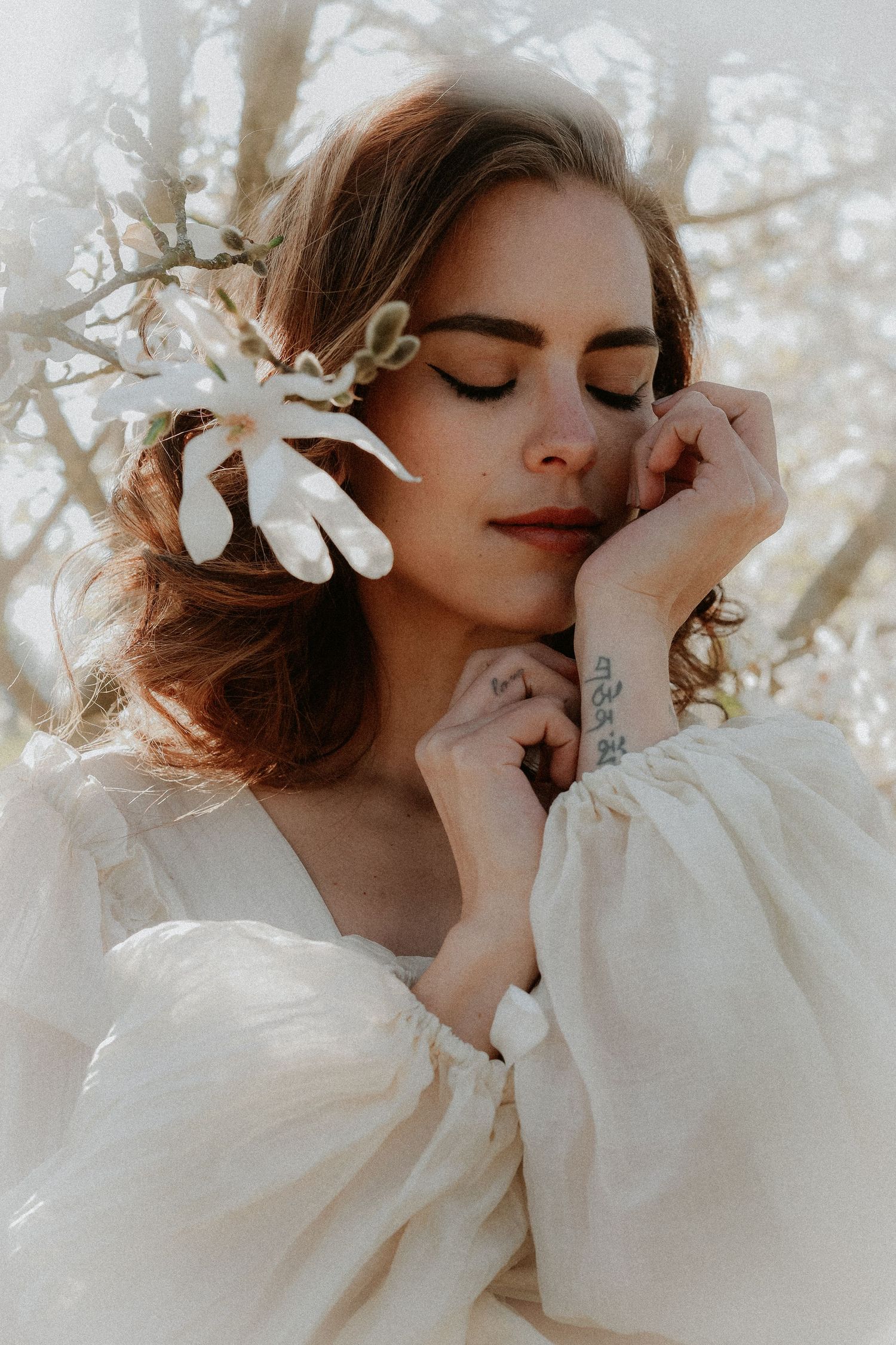 Woman with closed eyes, white flower in auburn hair, wearing flowing white dress, hands near face, soft dreamy outdoor setting.