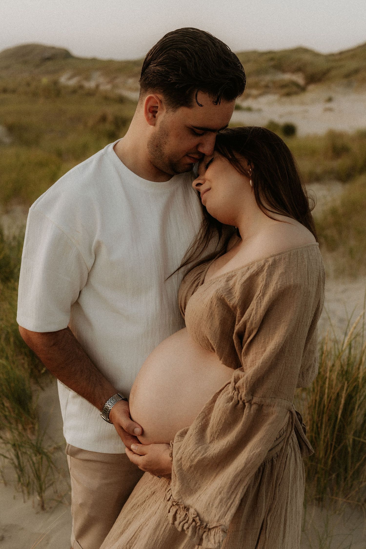 Couple embracing on sandy beach dunes at dusk, man kissing pregnant woman's forehead while holding her belly.