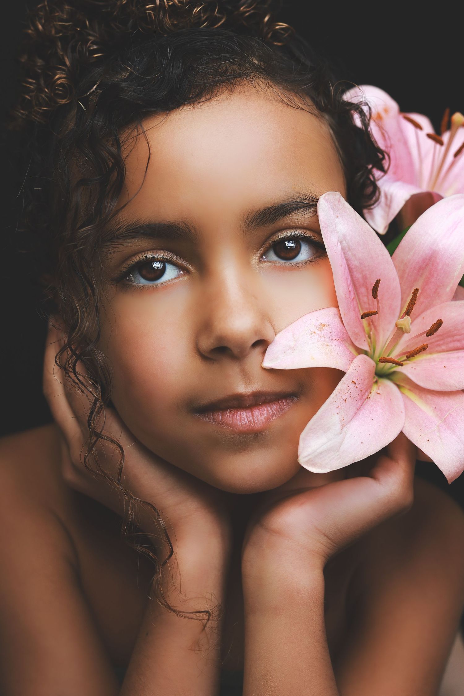 Young girl with curly hair resting chin on hands beside a pink stargazer lily flower.