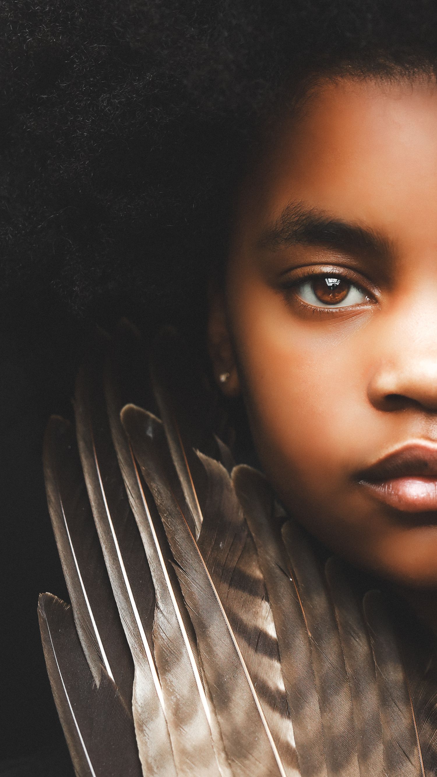 Close-up portrait of a young Black girl with natural afro hair holding feathers, gazing intensely against dark background.