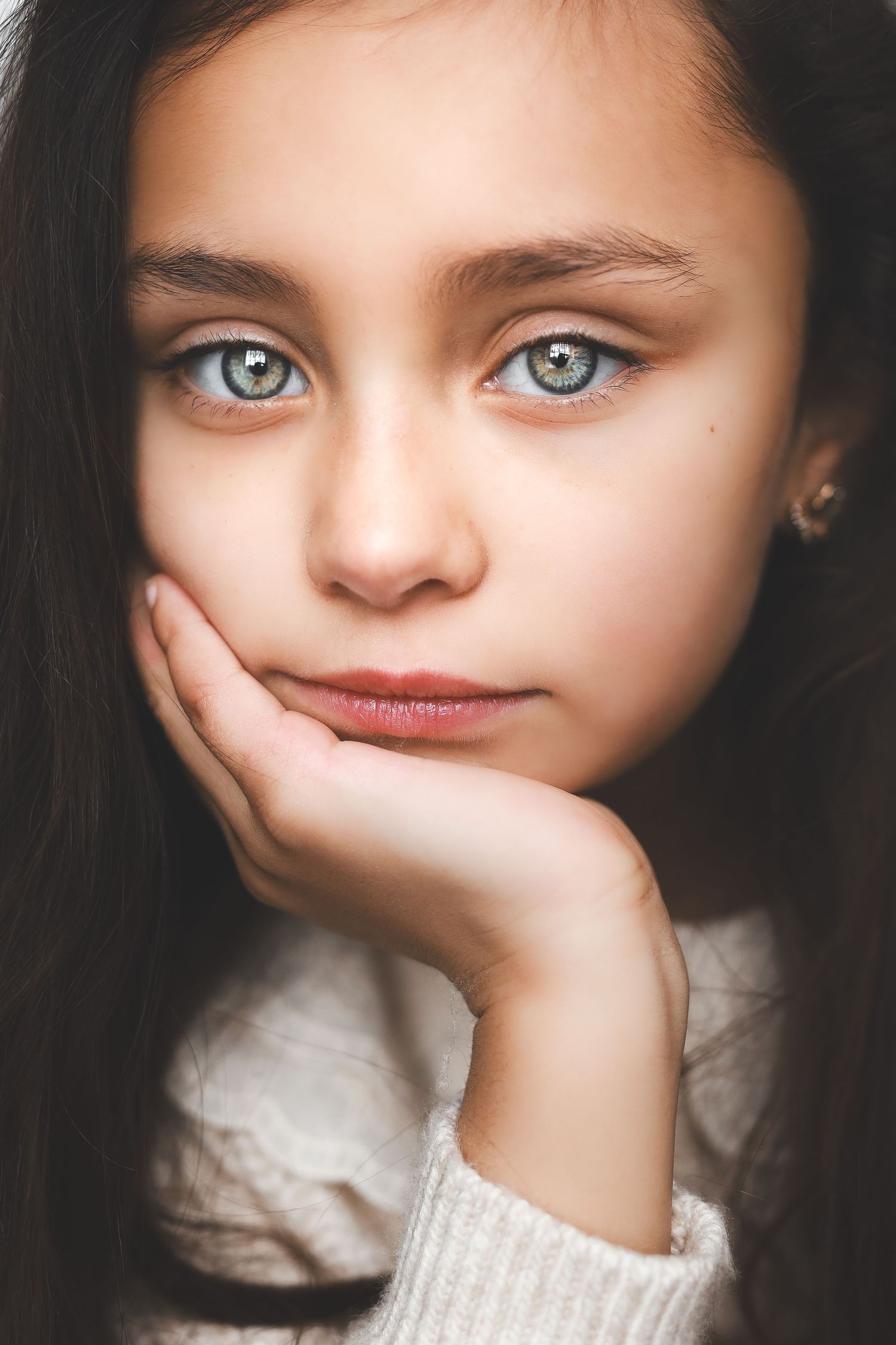 A young girl with striking blue-gray eyes rests her chin on her hand, gazing thoughtfully at the camera.