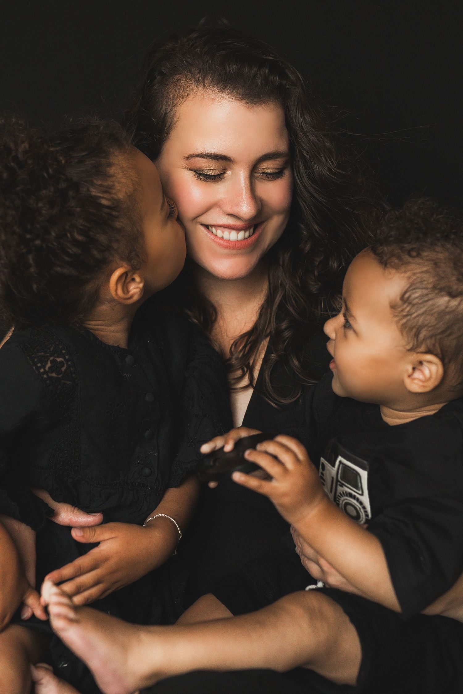 Smiling young mother with curly hair embraces her two young children against a dark background.