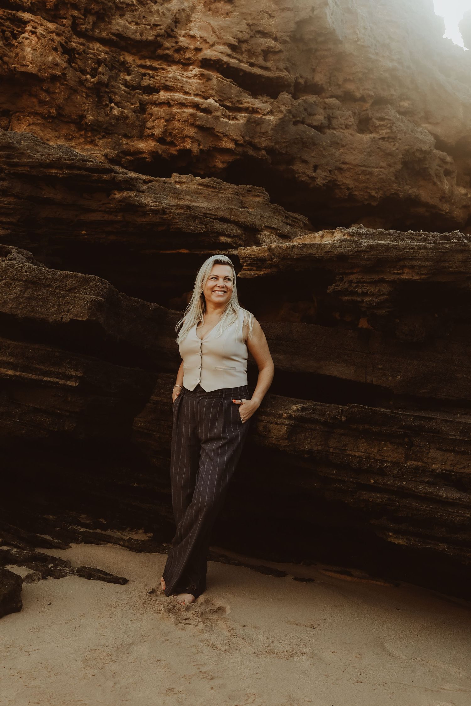 Smiling blonde woman in vest and wide-leg pants posing against sandstone rock wall.