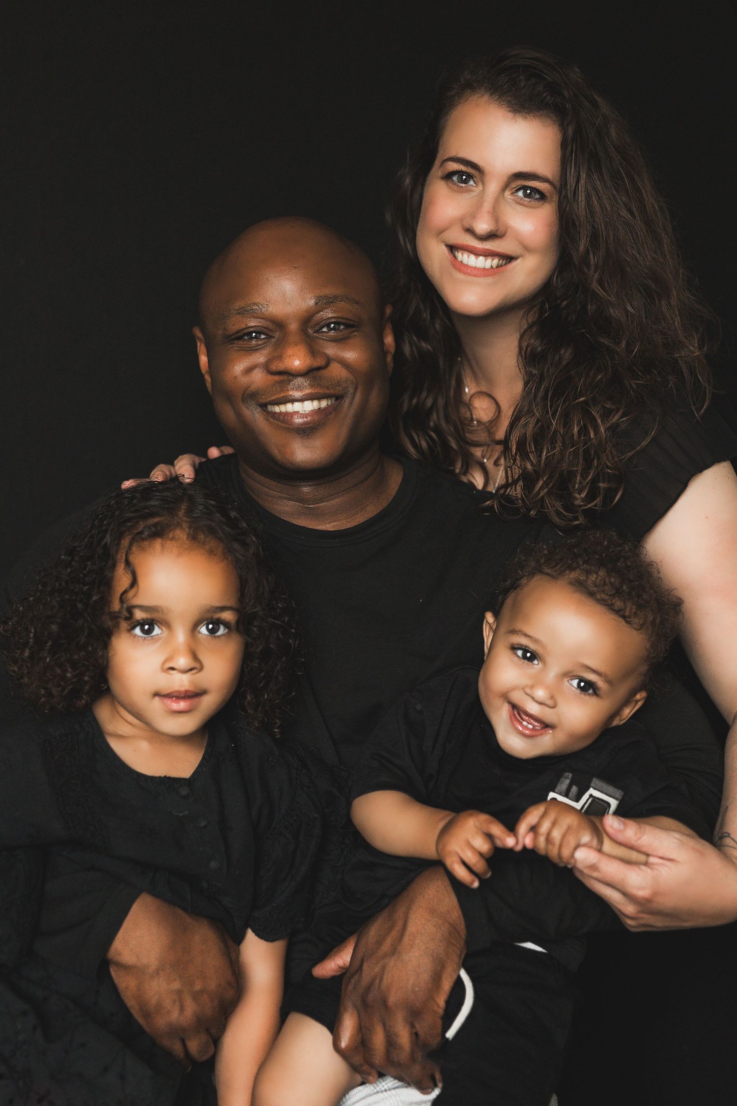 Smiling interracial family of four posing together in black outfits against dark background.