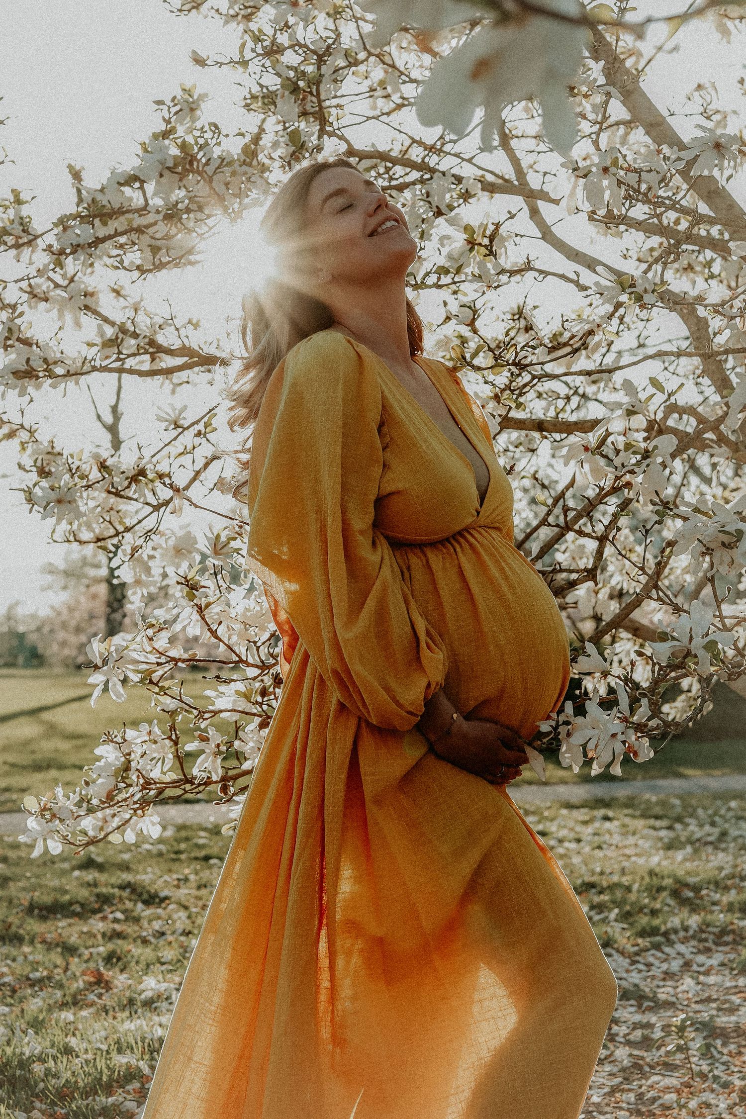Glowing pregnant woman in golden dress basking in sunlight beside flowering magnolia tree.