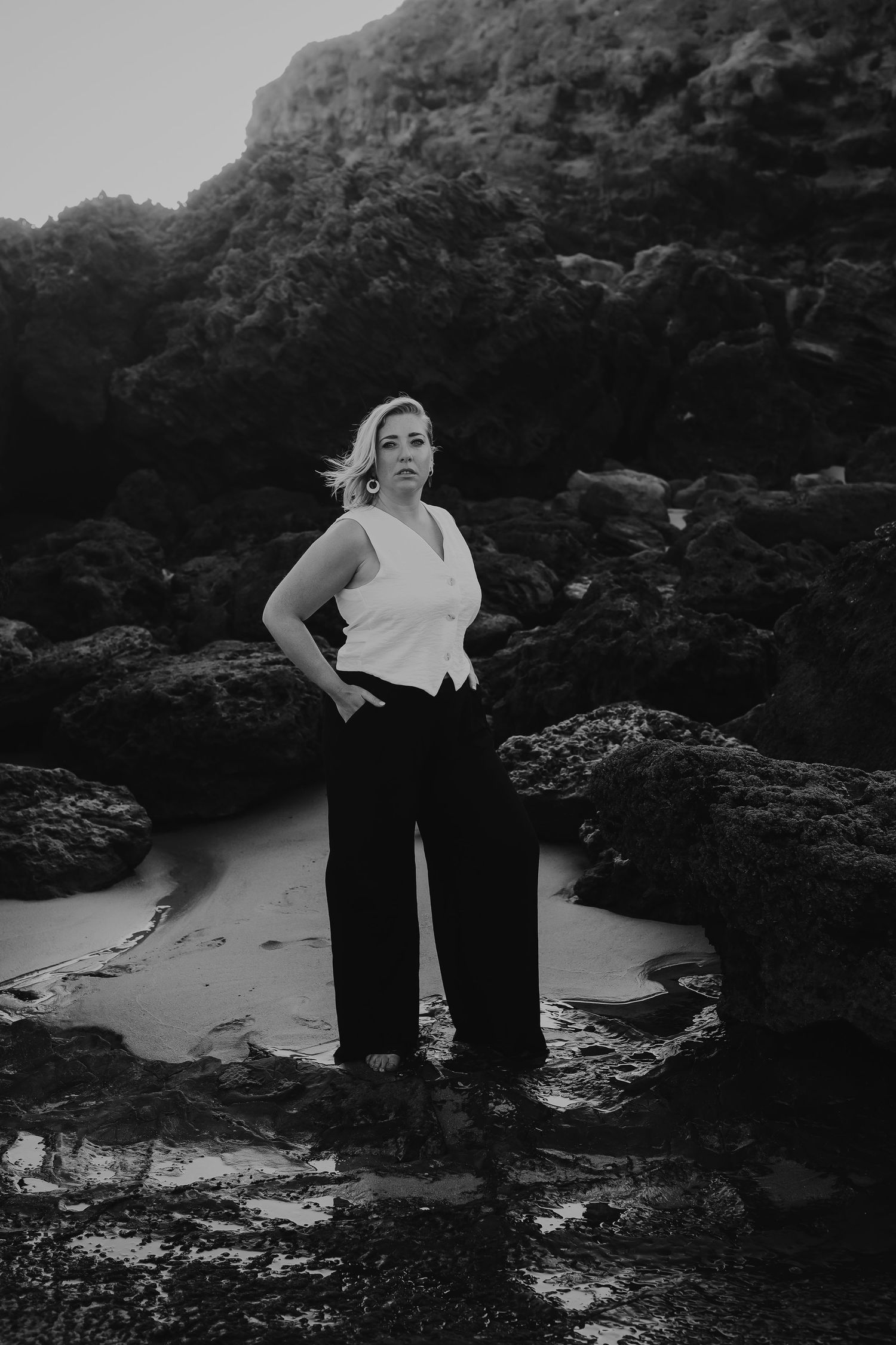 Black and white portrait of blonde woman in white top and black trousers standing on coastal rocks.