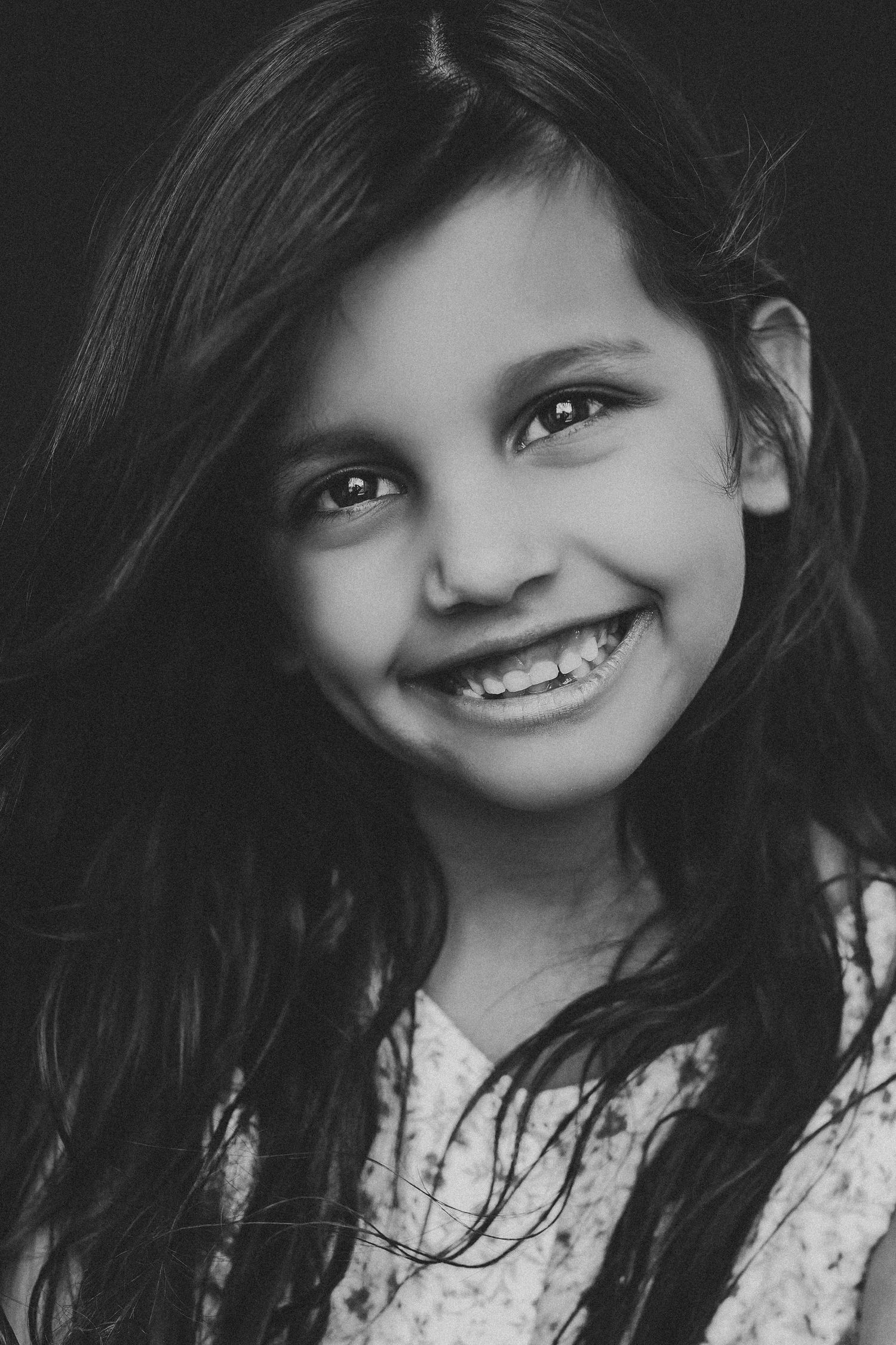 Black and white portrait of a smiling girl with long hair and braces against a dark background.