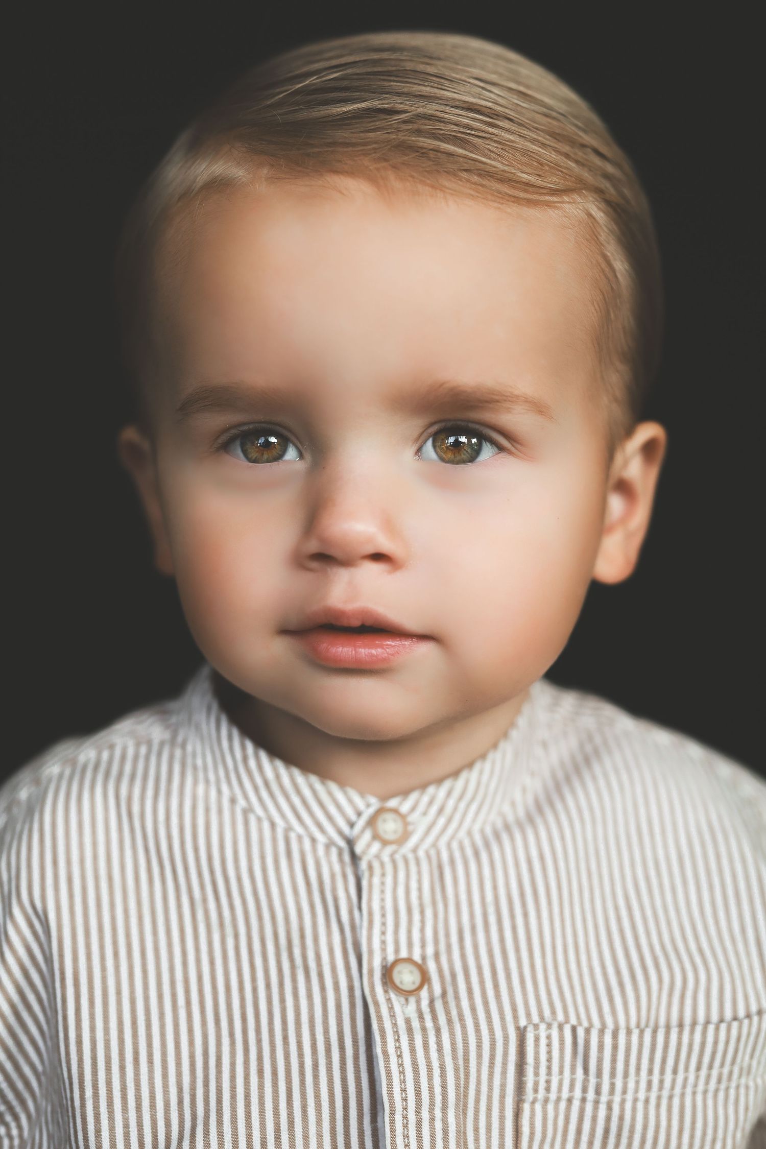 Portrait of a blonde toddler boy with brown eyes wearing a striped button-up shirt against dark background.