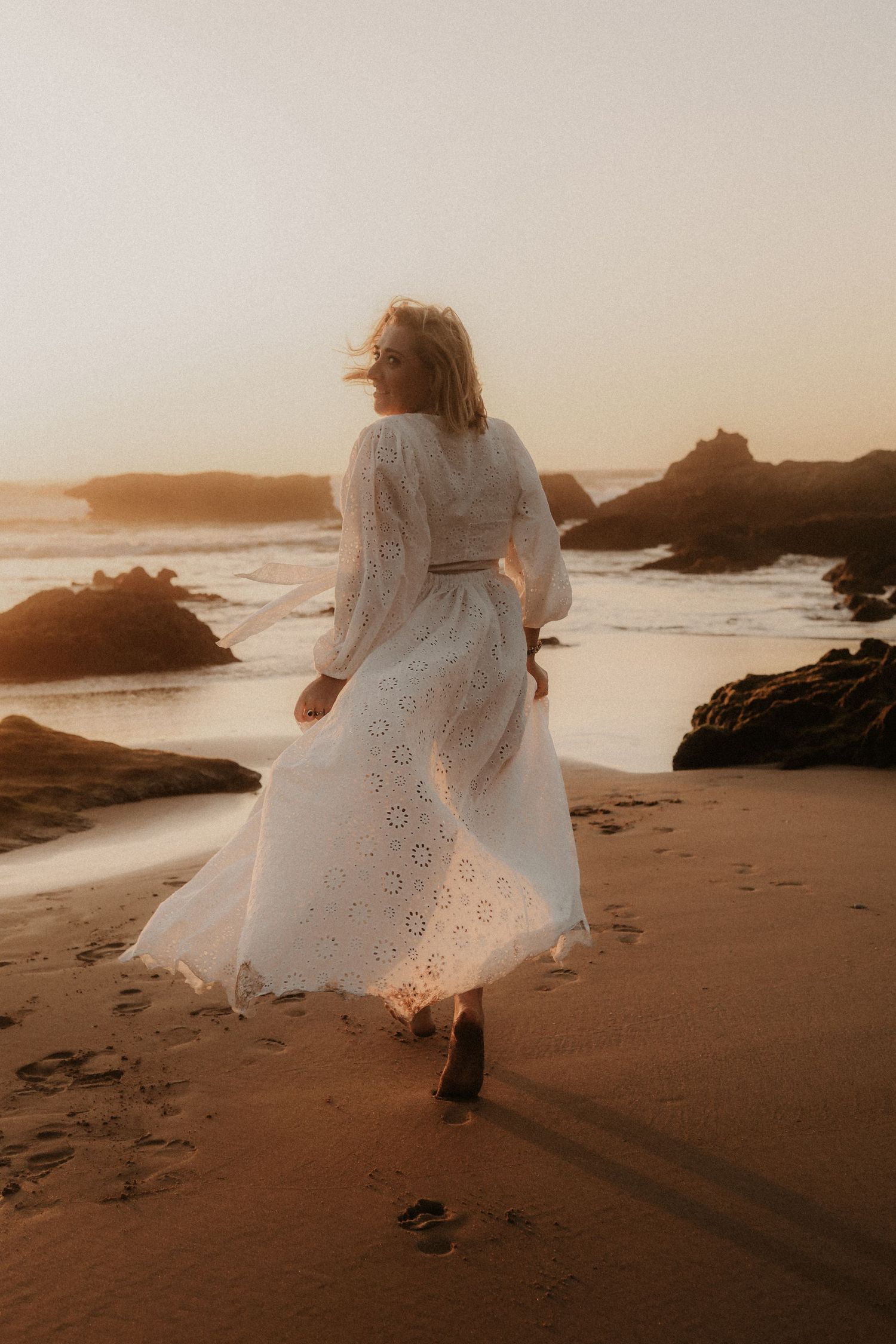 Woman in white lace midi dress walking on sandy beach during golden hour sunset.
