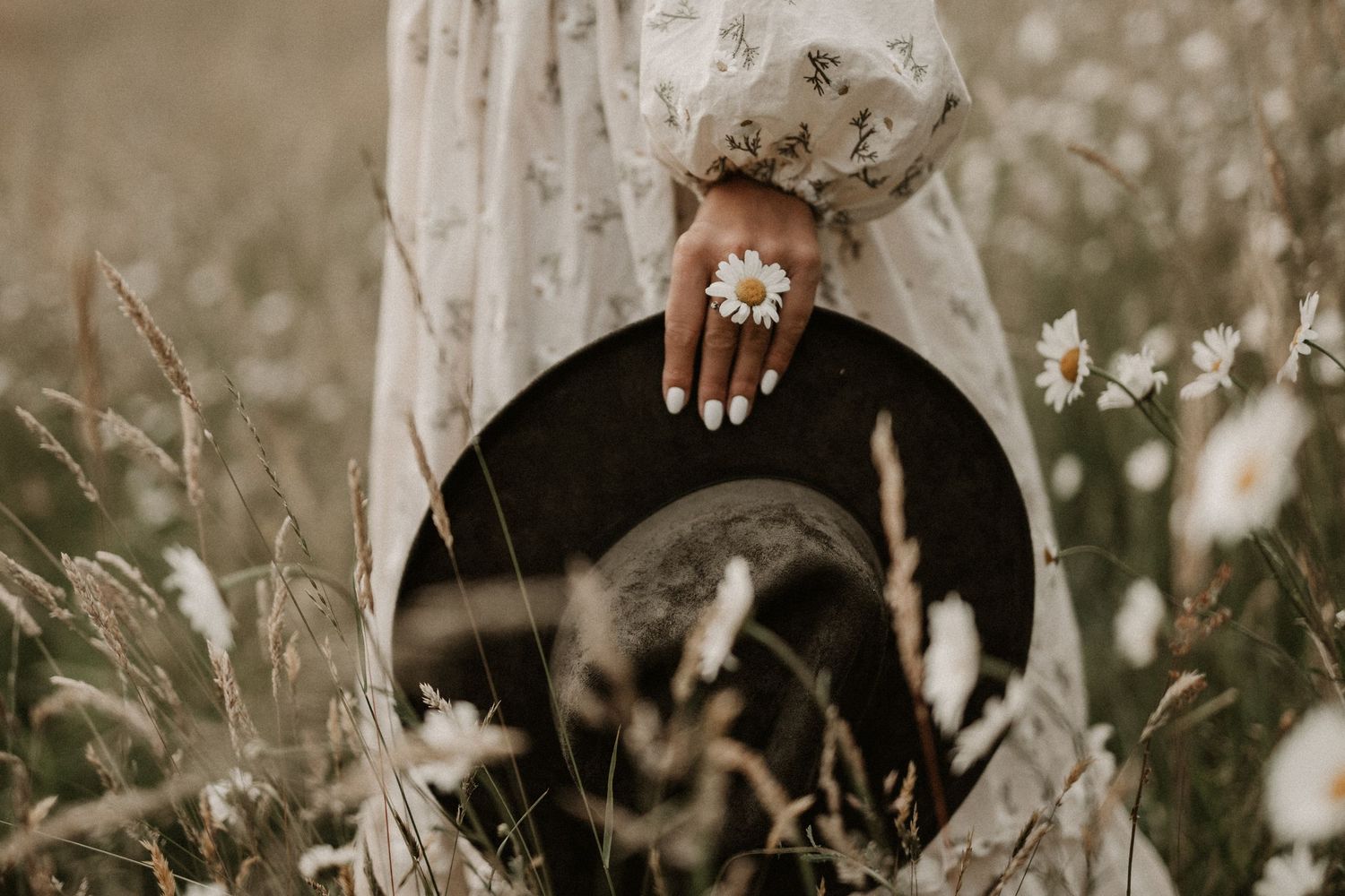 Close-up of a hand wearing a daisy ring holding a black hat in a wildflower field.