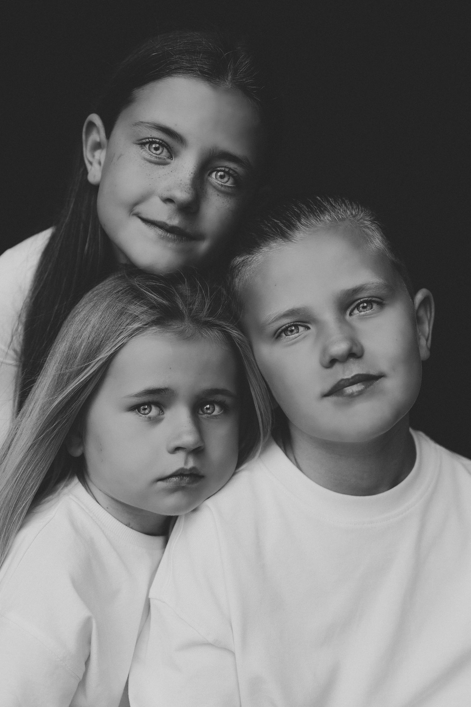 Black and white studio portrait of three siblings in white shirts against dark background.