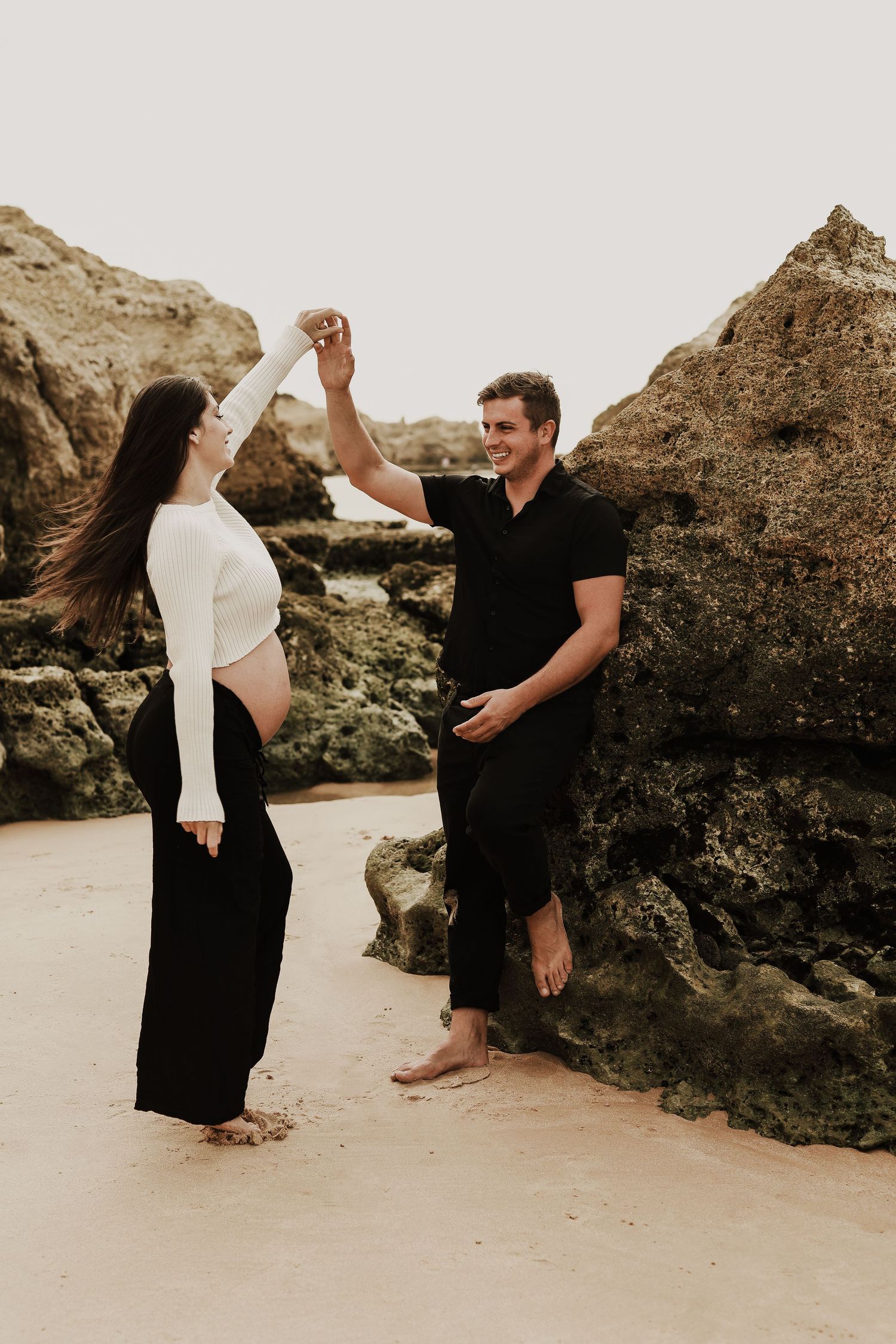 Happy couple dancing on sandy beach near large rocks, pregnant woman in white crop top.