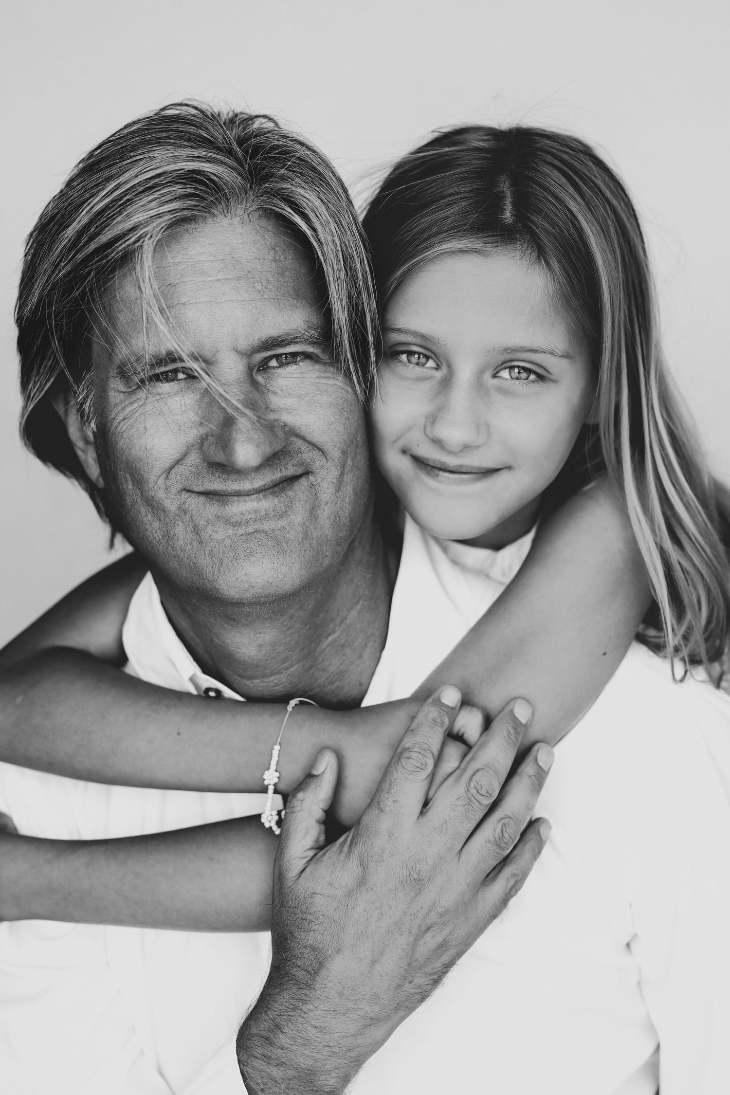 Black and white portrait of smiling father with young daughter hugging him from behind affectionately.