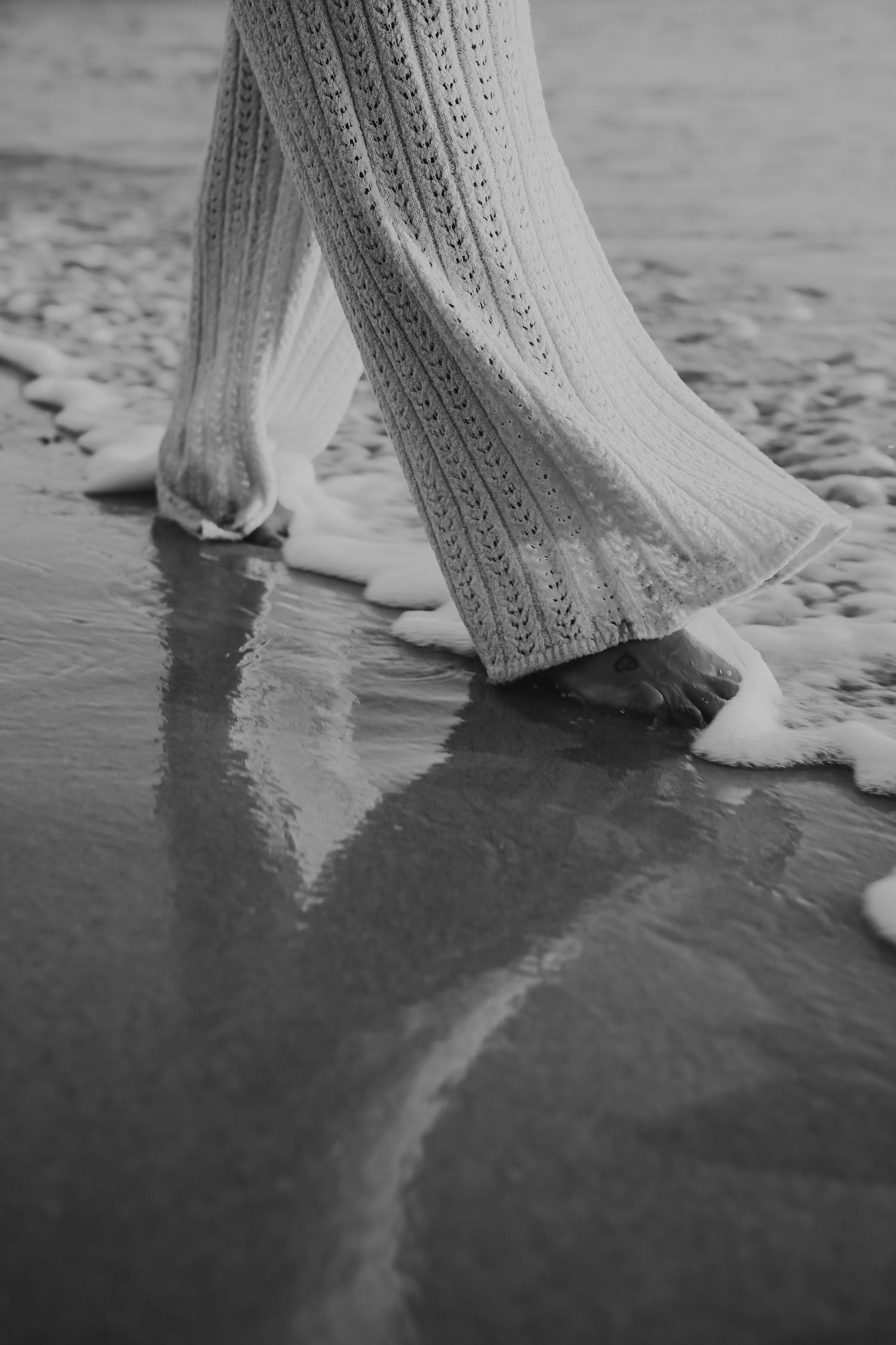Black and white close-up of bare feet walking through ocean waves in lace pants.
