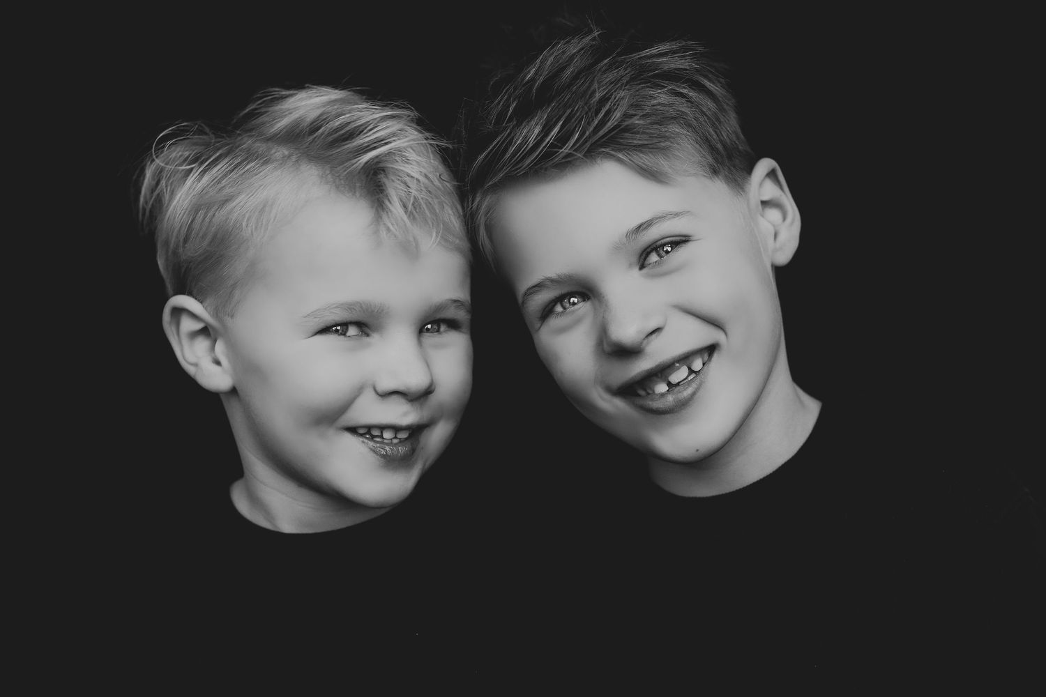 Black and white portrait of two cheerful young brothers in black shirts, smiling against dark background.