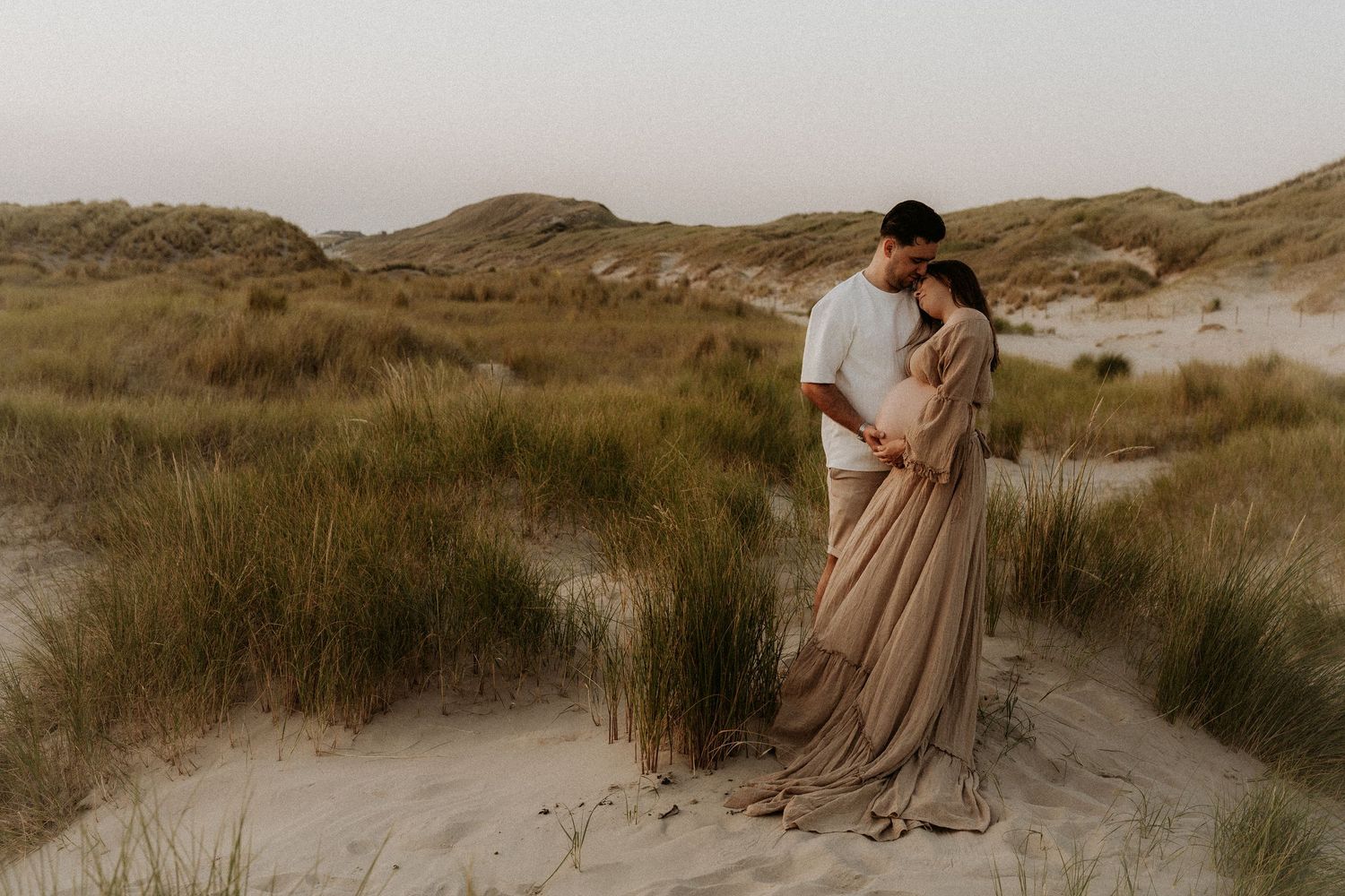 Couple embracing in sandy dunes at sunset during maternity photoshoot.