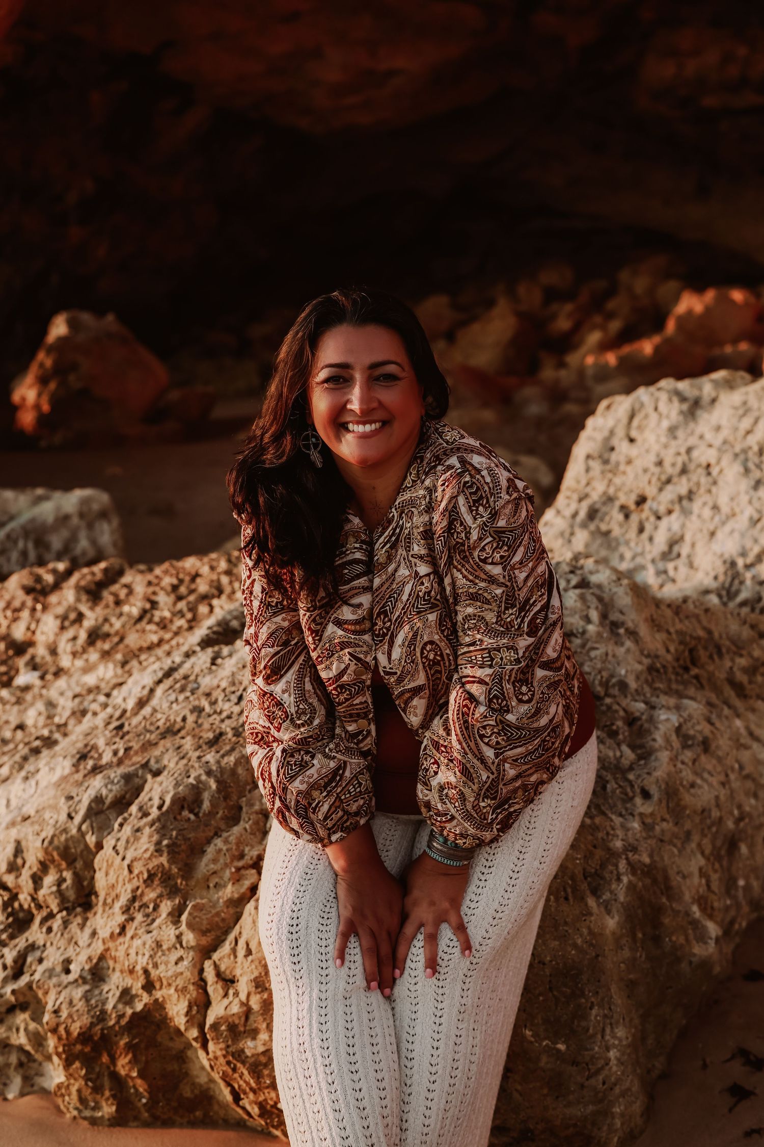 Smiling brunette woman in paisley blouse posing against rocky cave backdrop.