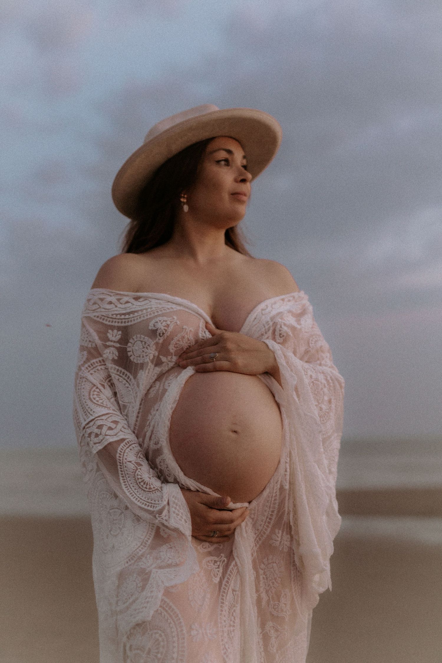 Pregnant woman in white lace off-shoulder dress and hat holding baby bump on cloudy beach.