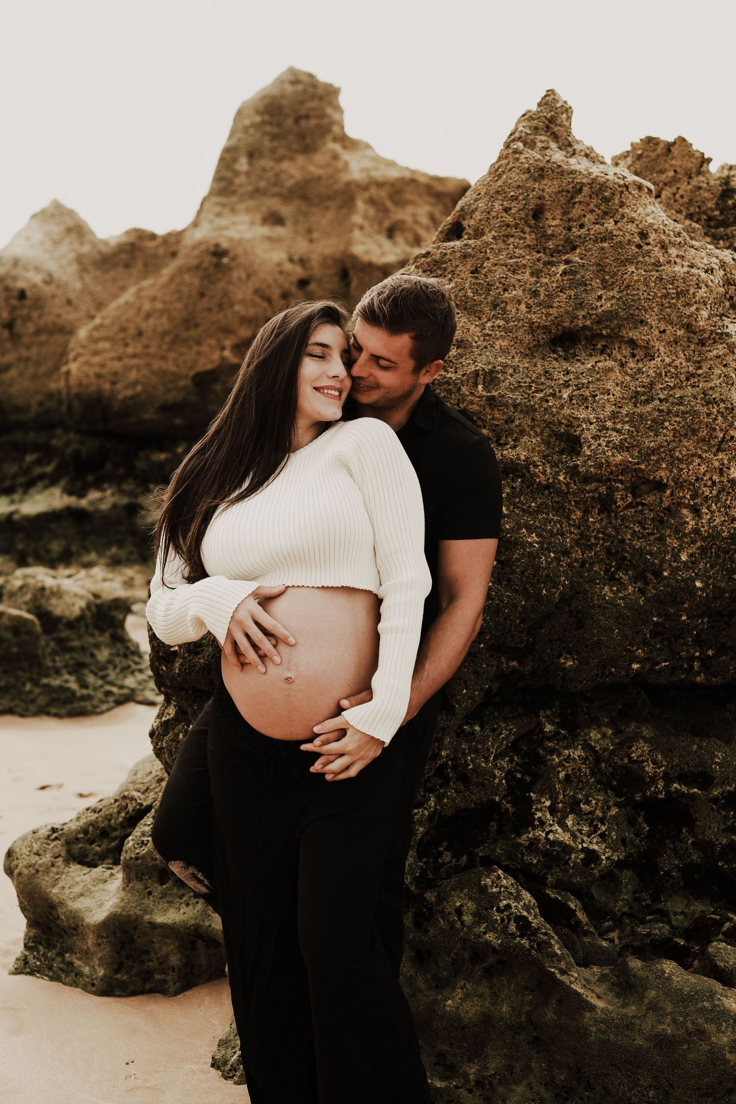Loving couple embracing on beach, man kissing pregnant woman near large rocky formation.