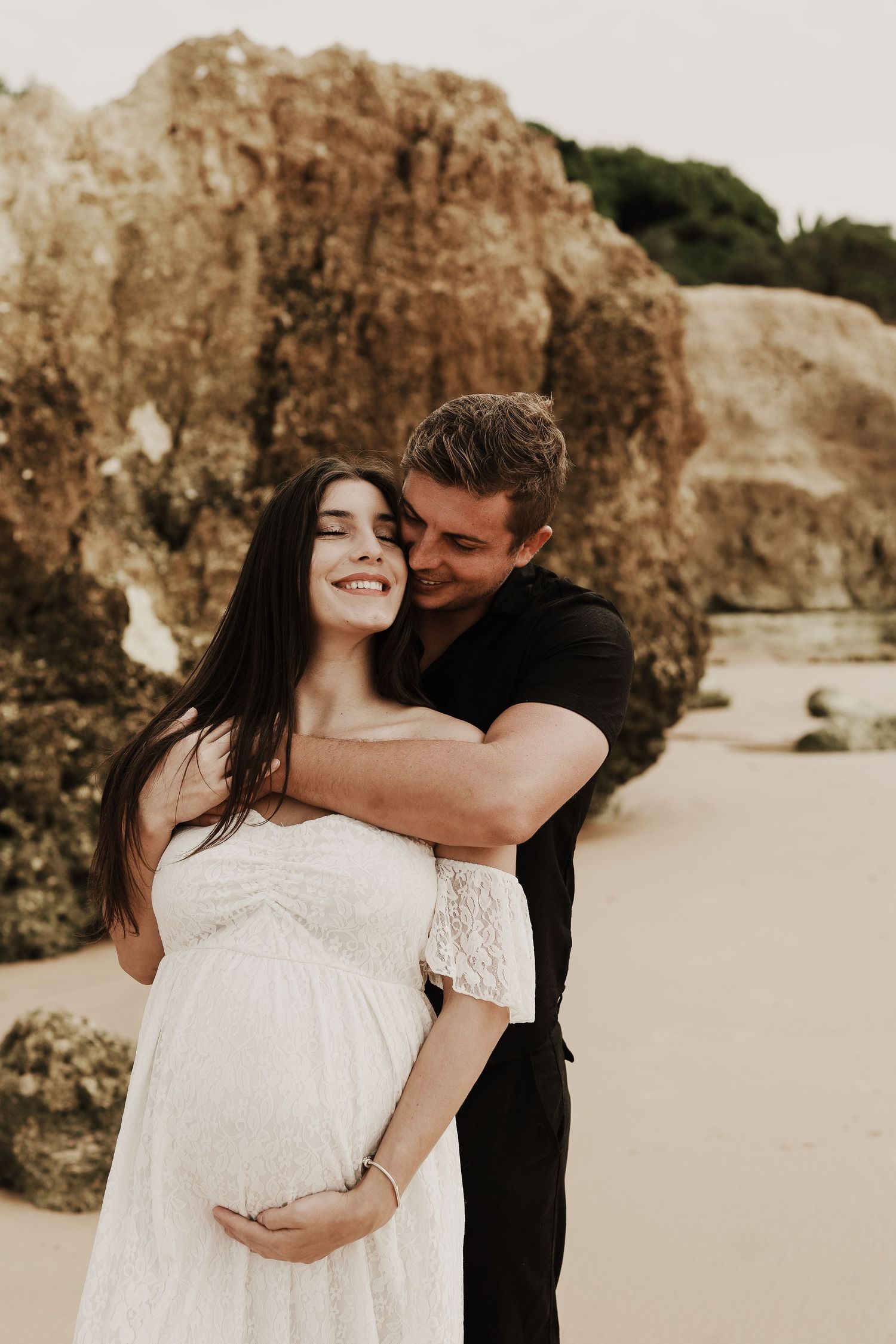 Couple embracing on beach, man kissing pregnant woman in white lace off-shoulder maternity dress.