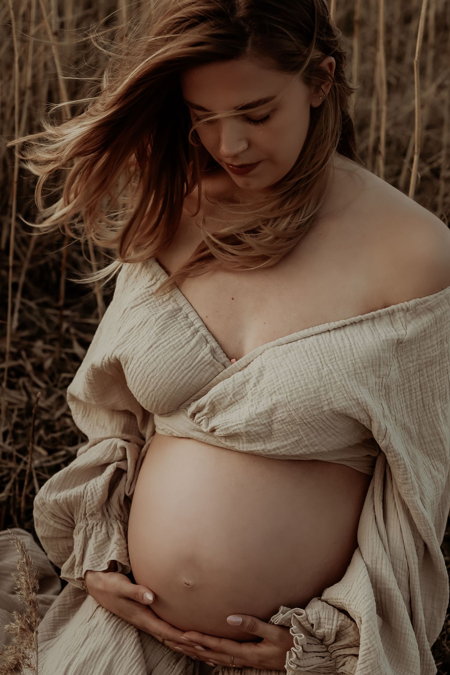 Pregnant woman with windswept hair wearing open linen wrap top cradling bare baby bump in field.
