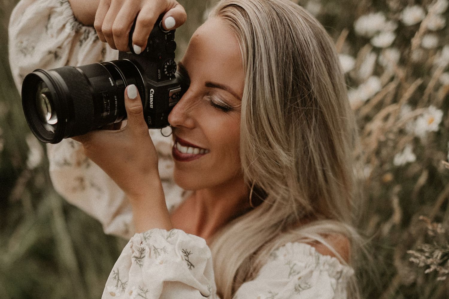Smiling blonde woman photographer holding a Canon camera outdoors in a wildflower field.