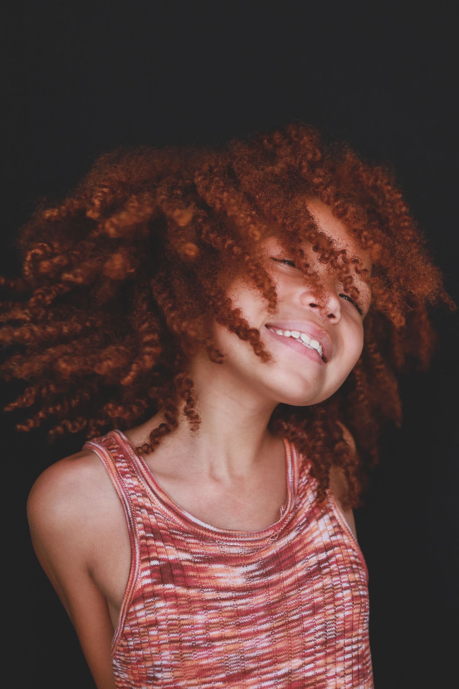Young woman with vibrant red curly hair tossing her head, smiling joyfully against a dark background.
