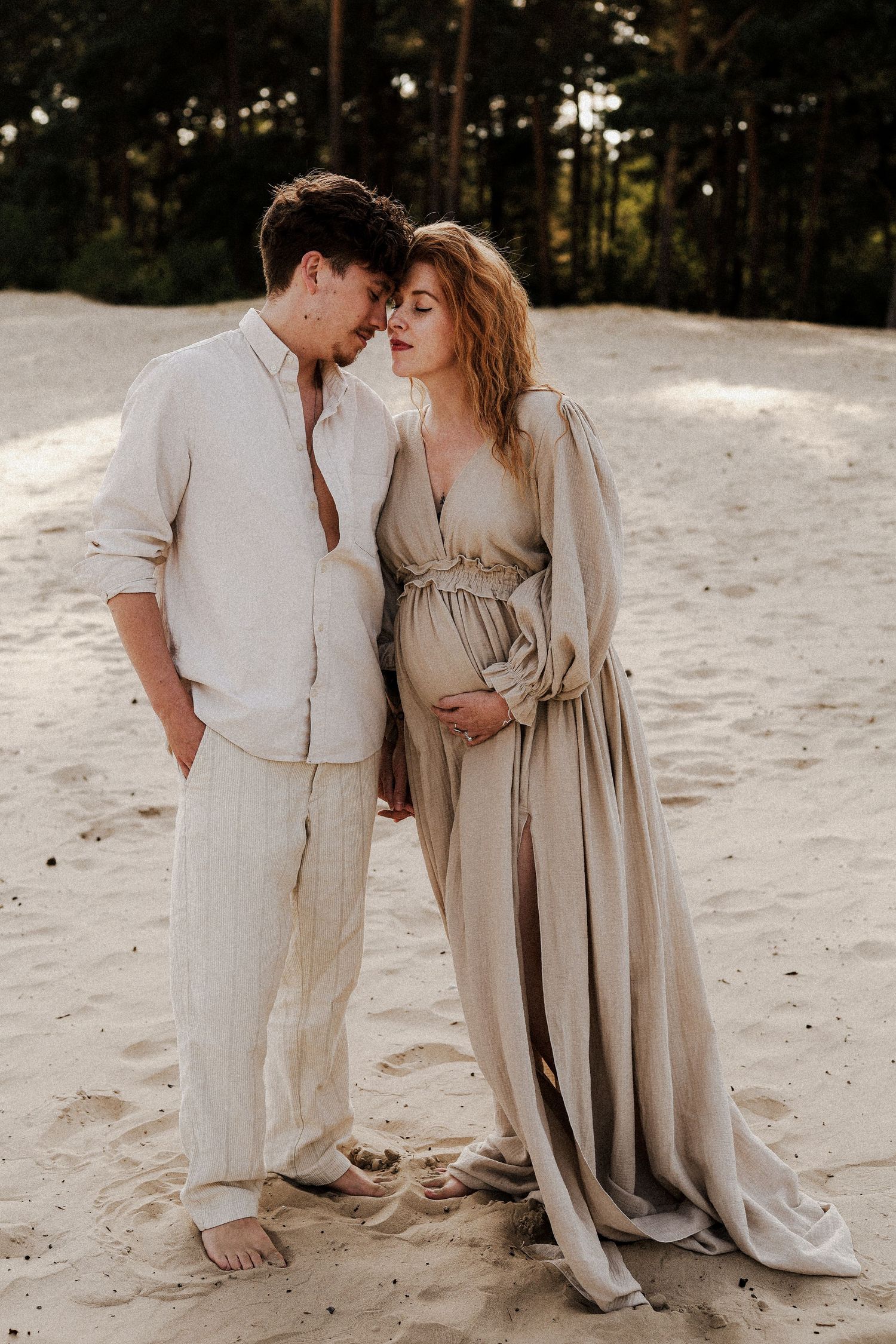 Elegant couple standing on sandy dunes, pregnant woman in flowing beige maternity dress.