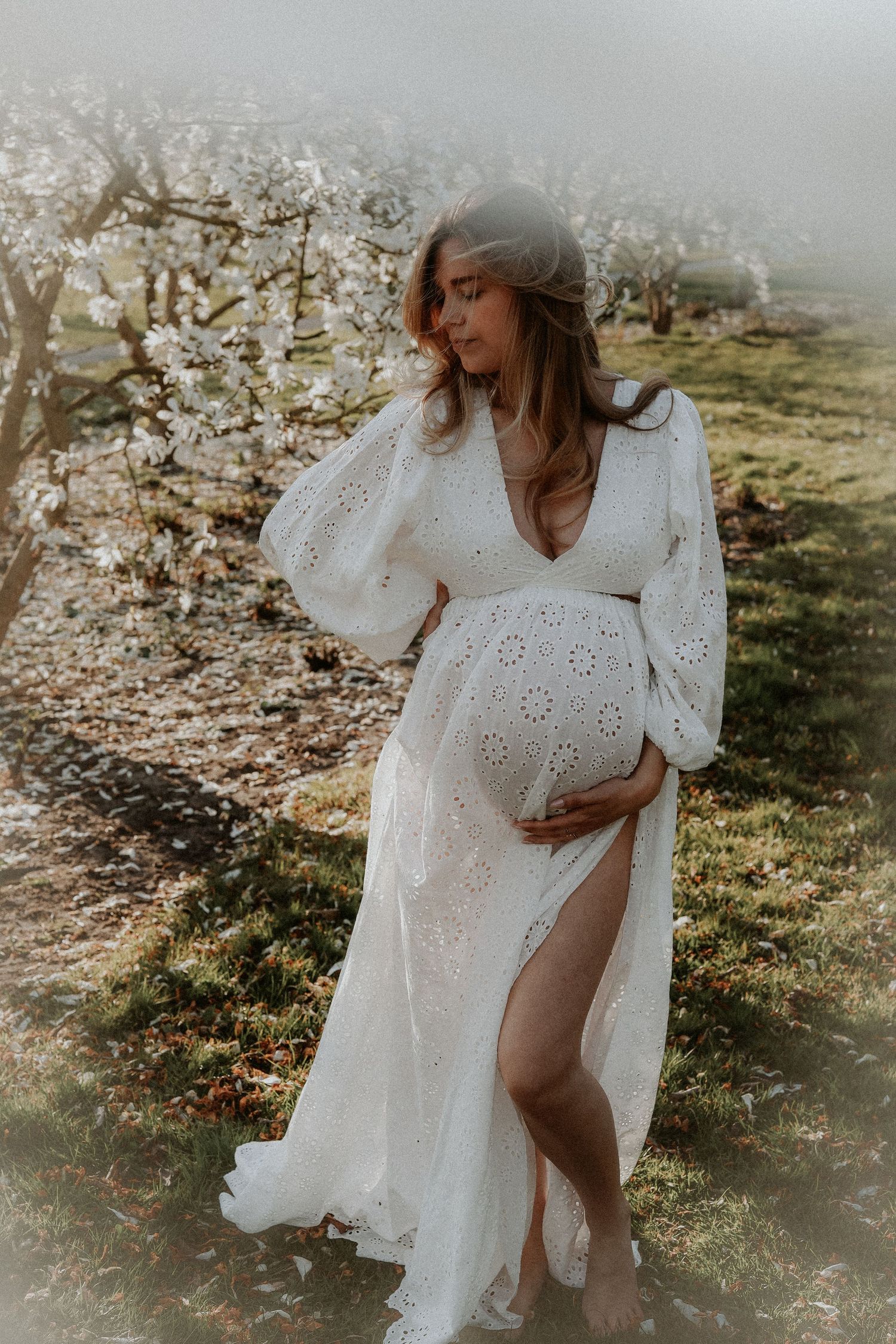 Pregnant woman in elegant white lace dress standing in misty blooming orchard garden.