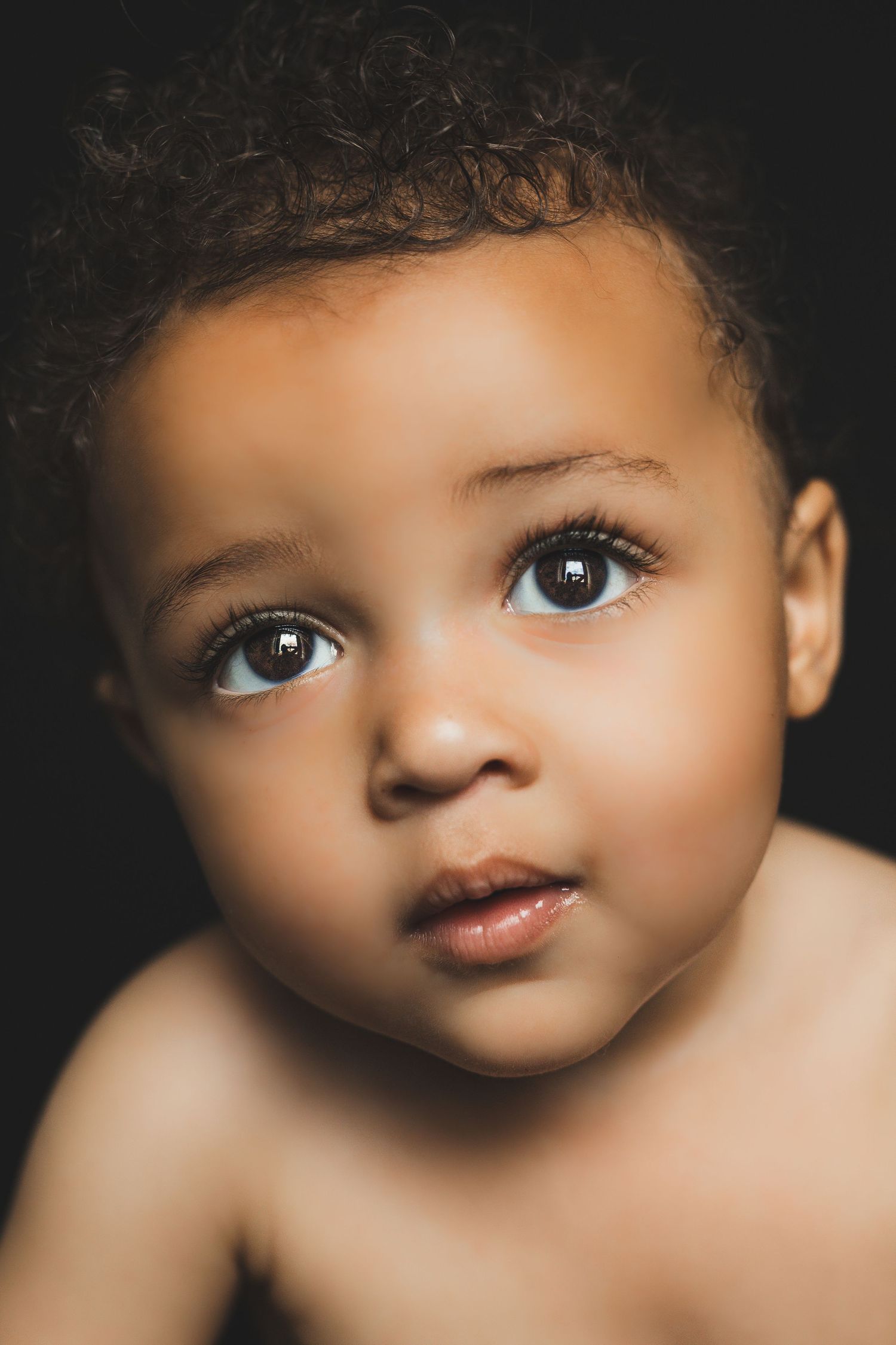 Close-up portrait of a cute baby with curly hair and expressive brown eyes on dark background.