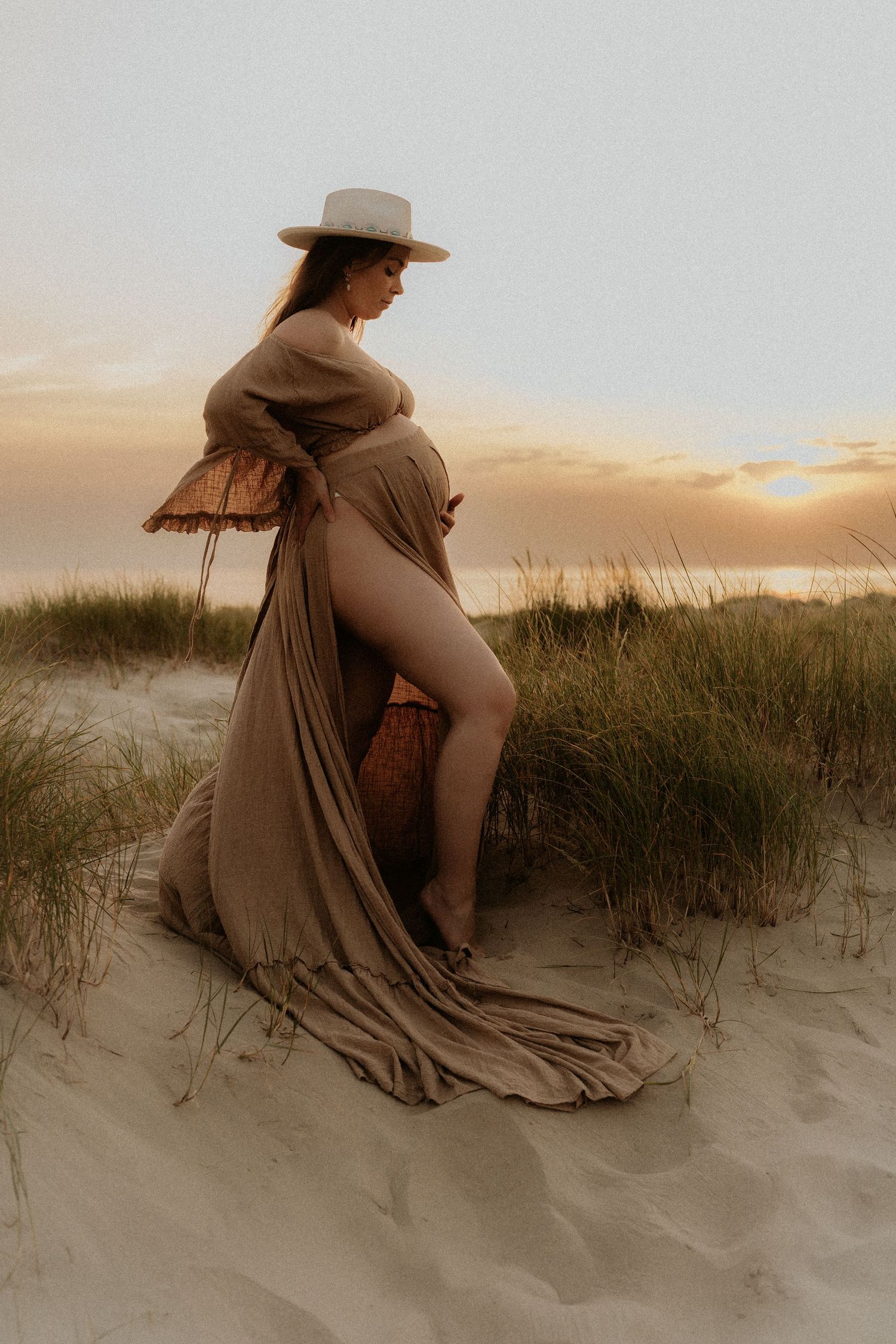 Pregnant woman in flowing brown wrap dress and wide-brim hat standing on beach dunes at golden sunset.