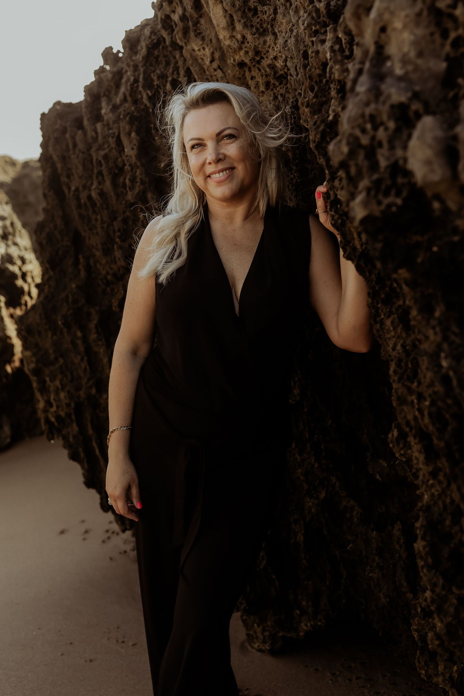 Smiling blonde woman in black jumpsuit posing between dramatic rocky cliffs on a beach.