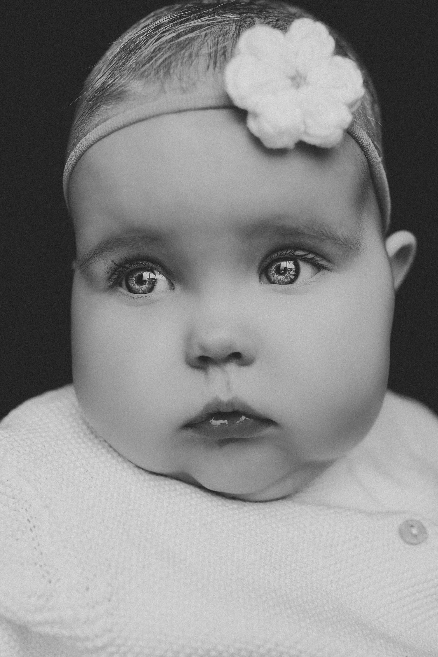 Black and white portrait of a baby girl wearing a white flower headband with big expressive eyes.