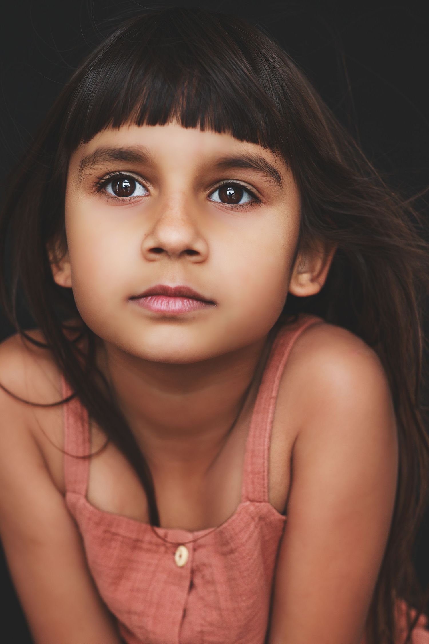 Portrait of a young girl with dark hair and bangs, gazing intensely at the camera against a dark background.