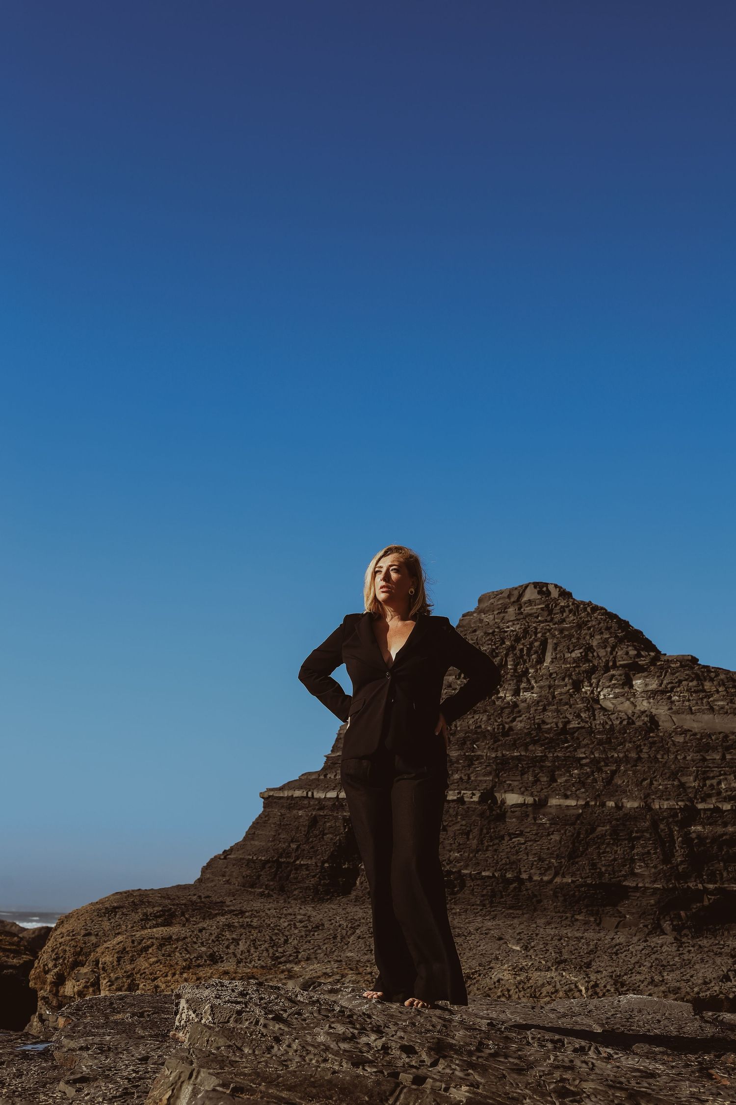 Blonde woman in black suit standing confidently on rocky coastal cliffs against a clear blue sky.