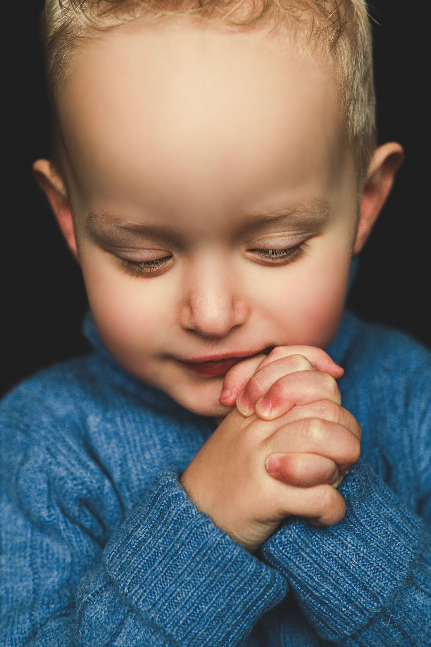 Close-up of a young toddler boy with blonde hair praying with hands clasped, wearing a blue knit sweater.