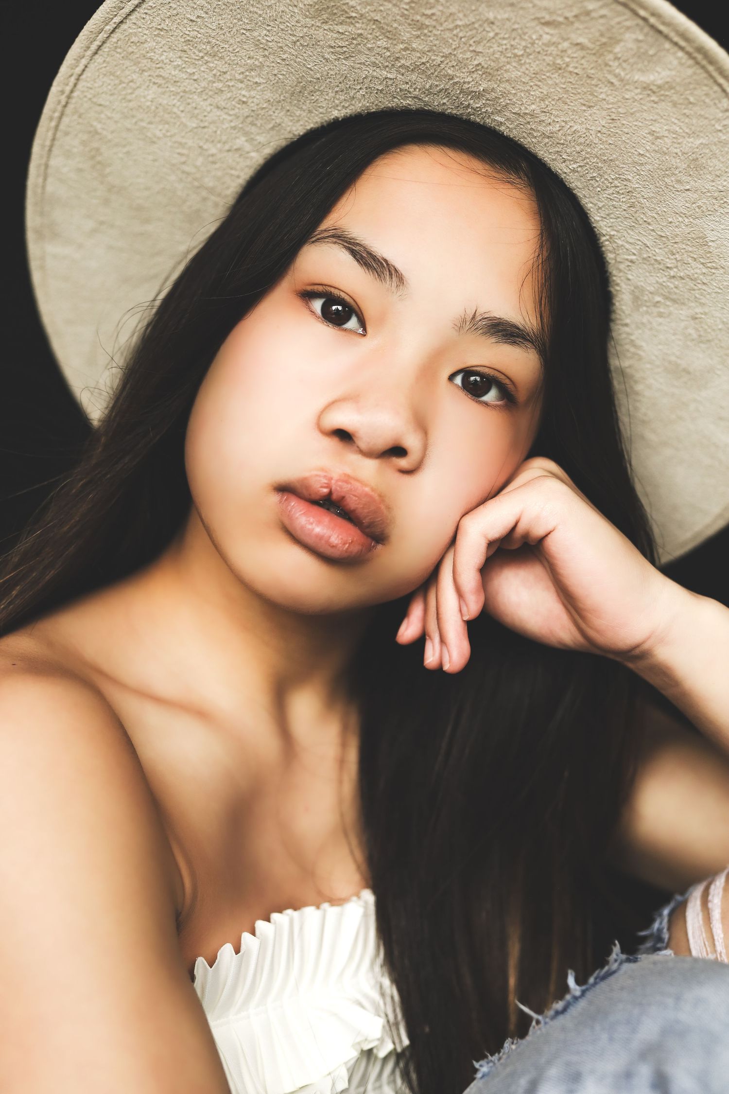 Young Asian woman in a wide-brim hat and white top, resting chin on hand, gazing at camera.