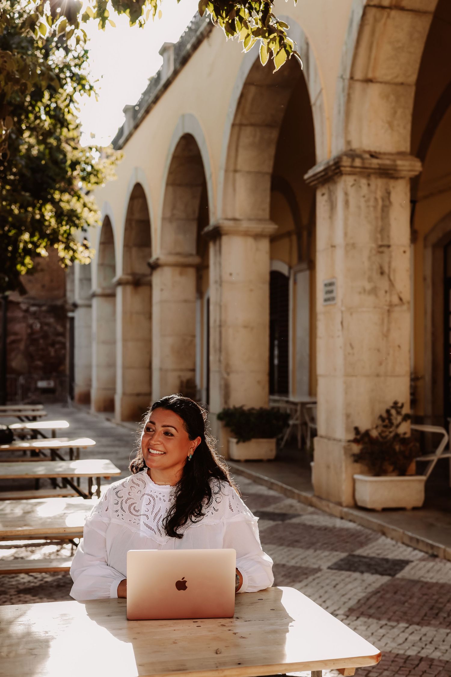 Smiling woman with laptop at outdoor table beneath arched stone building in sunlight.