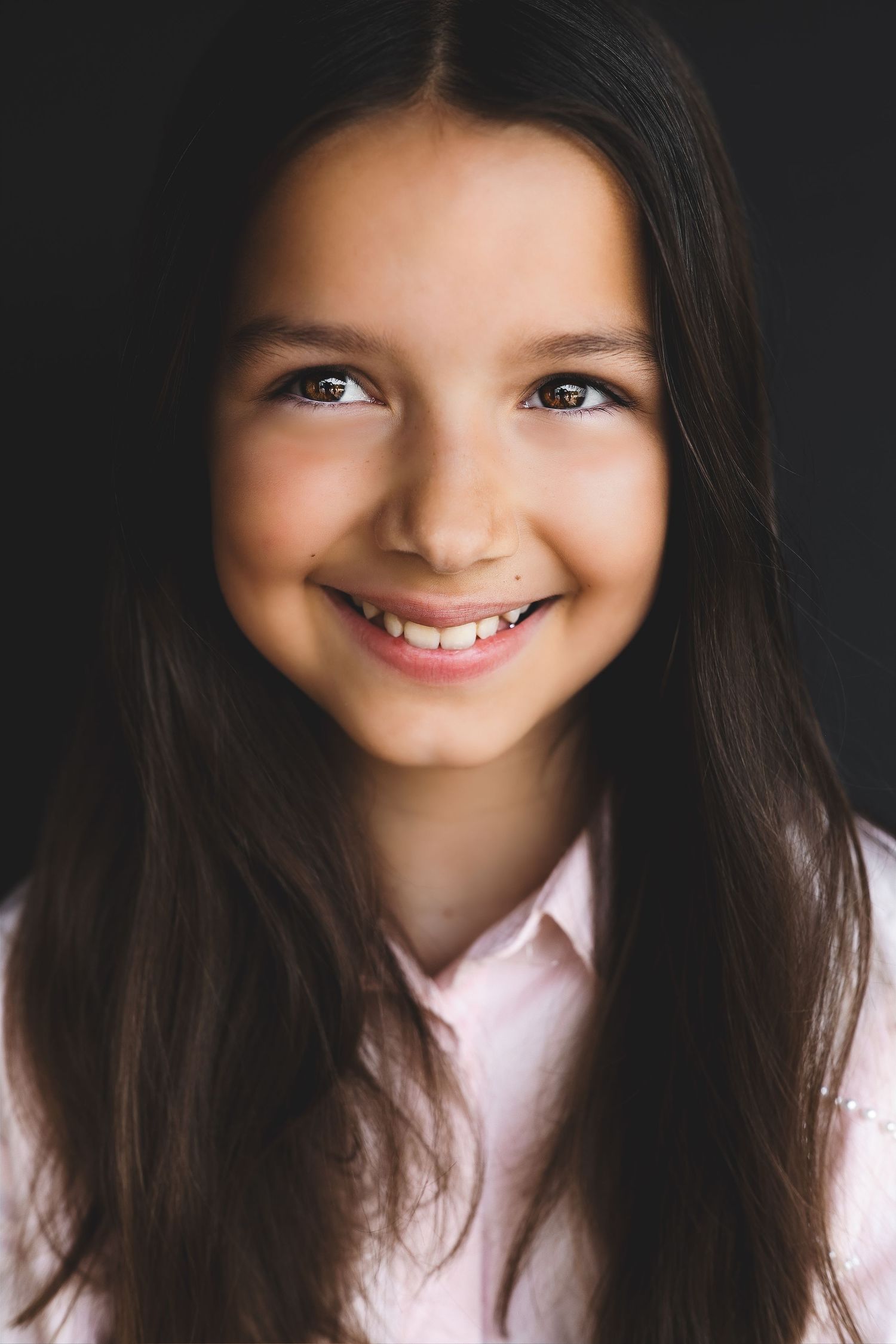 Smiling young girl with long dark hair wearing a pink shirt, portrait photo on dark background.