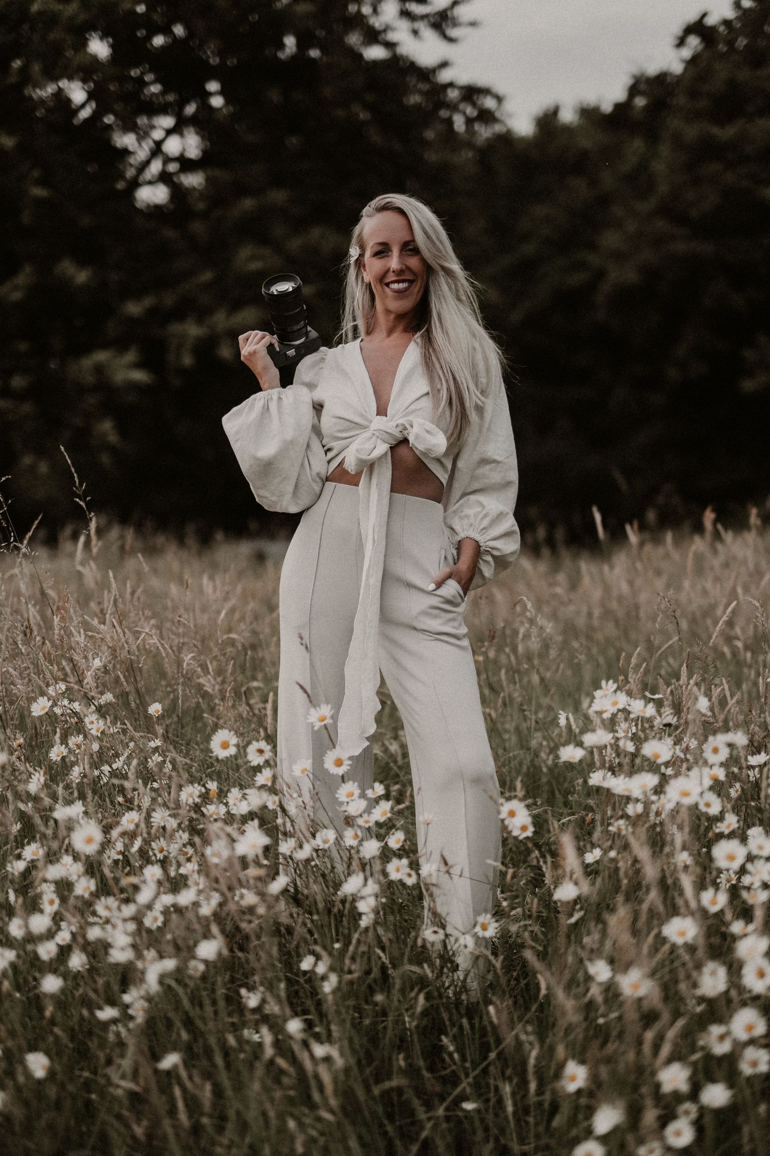 Smiling blonde woman in white co-ord outfit holding a camera in a field of white wildflowers.