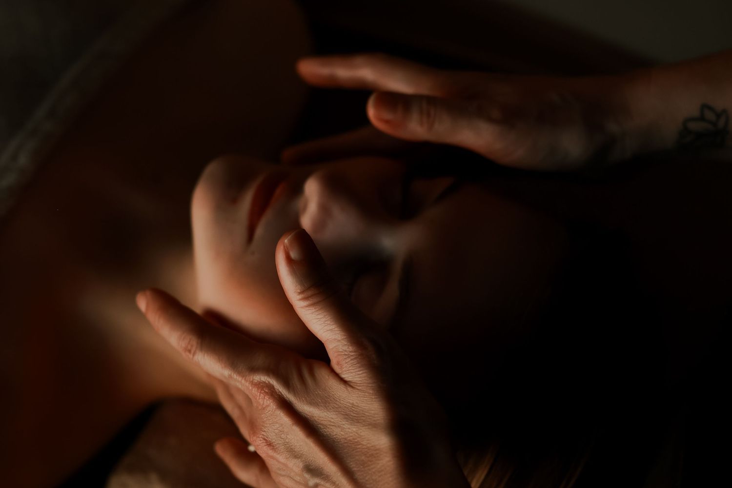 Close-up of hands with tattoos gently cradling a person's face in dramatic low-key lighting.