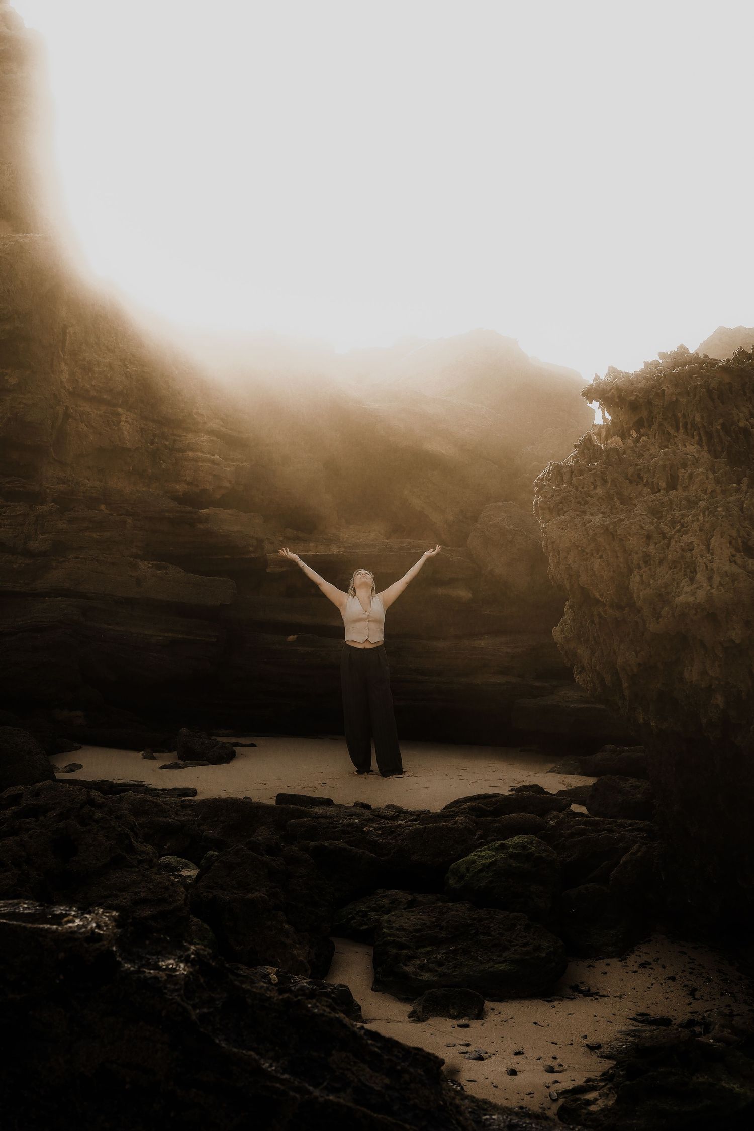 Woman standing with arms raised in misty rocky cave with dramatic light rays.