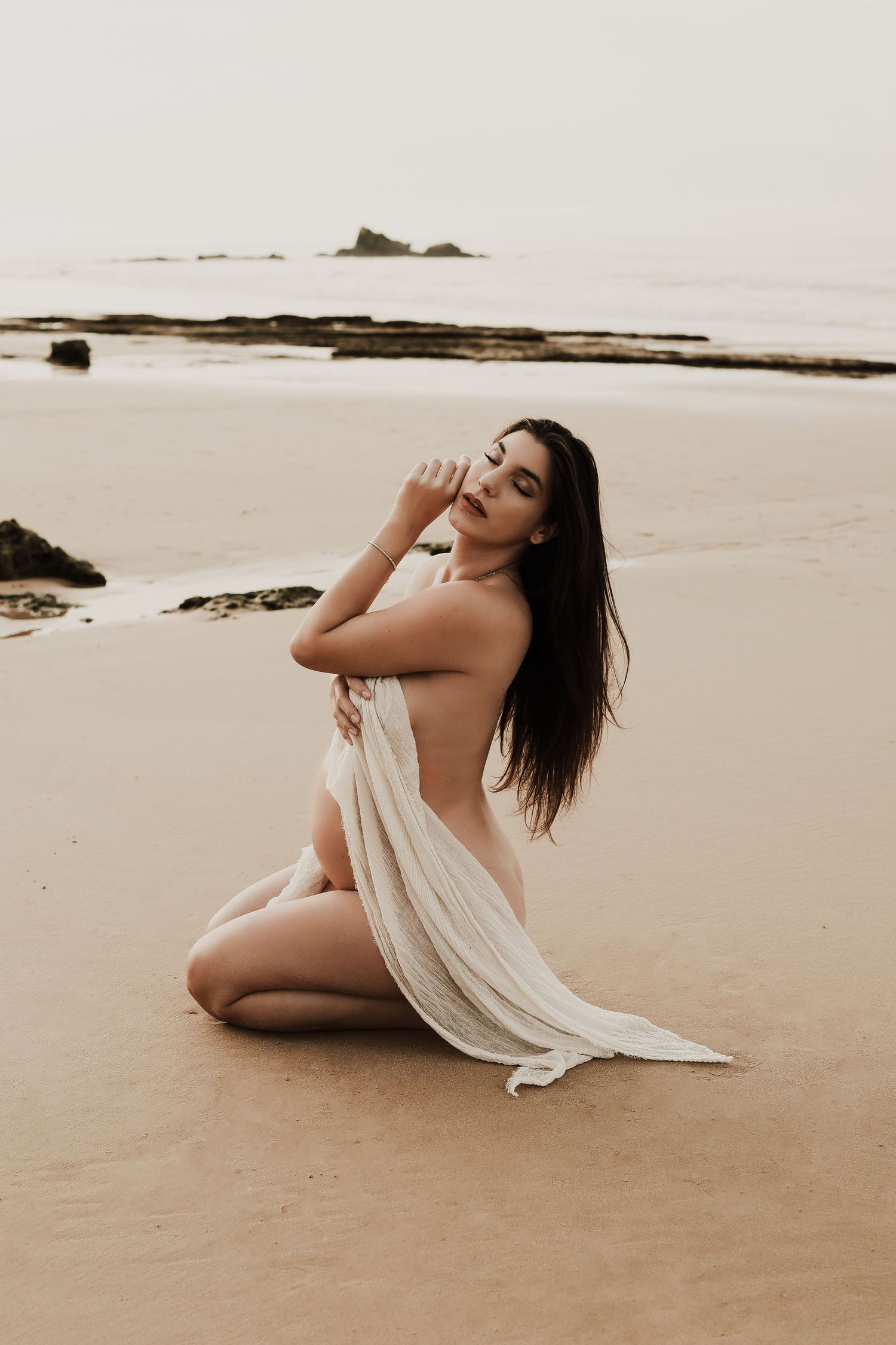 Woman wrapped in white fabric sitting gracefully on sandy beach with ocean in background.